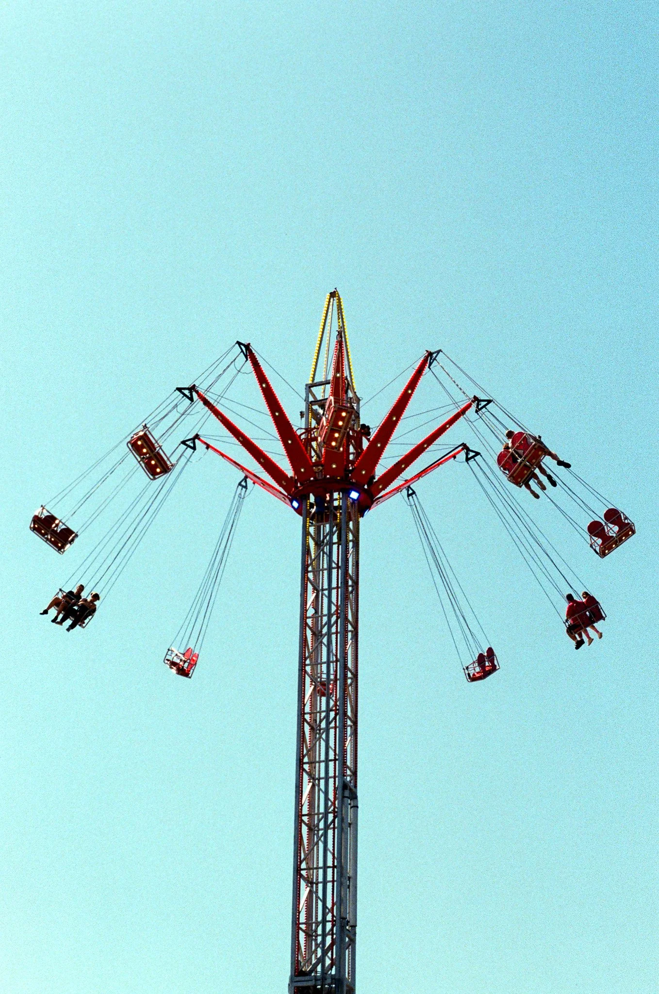 A tall amusement park ride with swinging chairs, some holding riders, against a light blue sky.