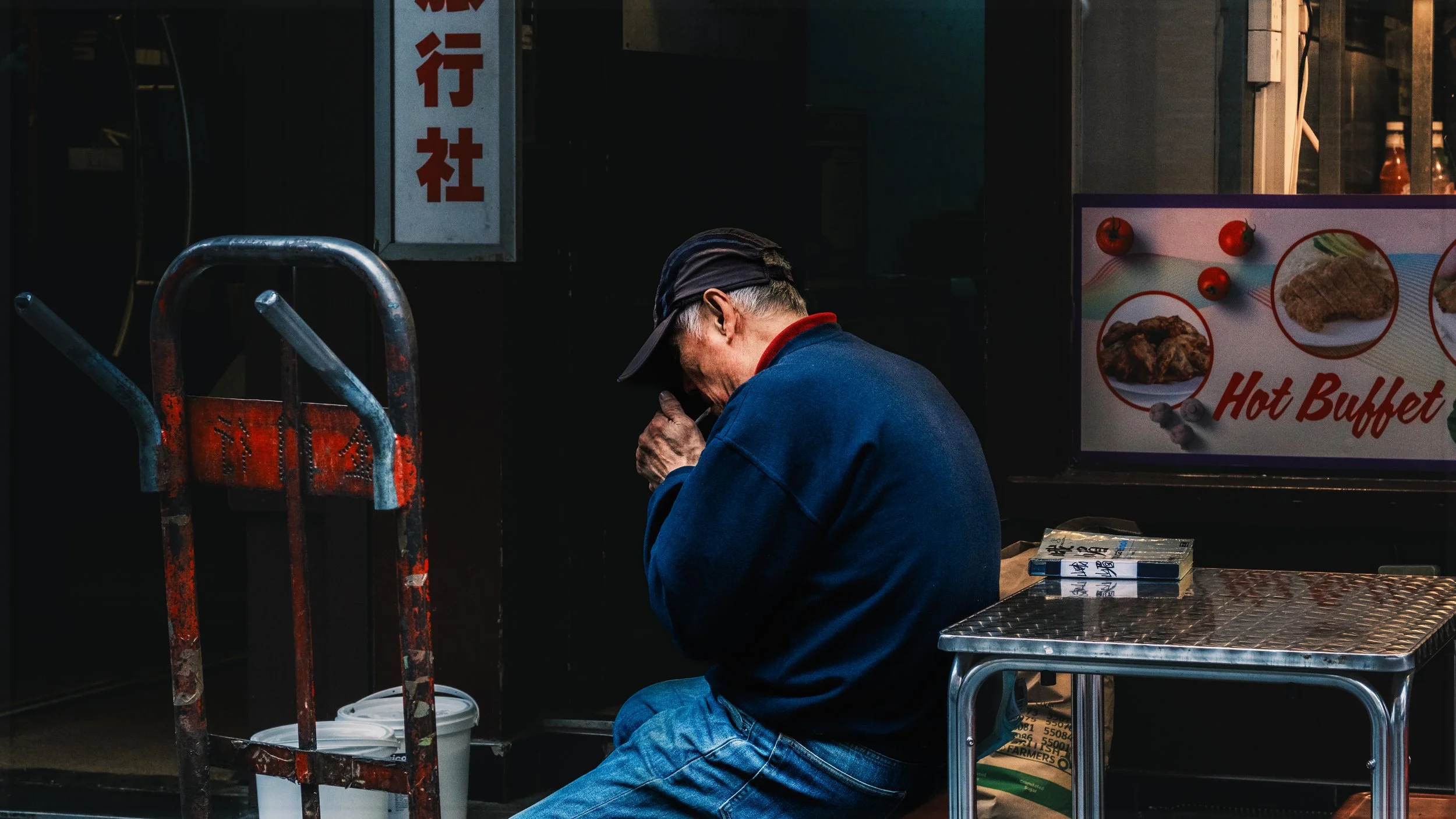 An elderly man with gray hair, wearing a dark cap and blue jacket, crouches down and appears to be lighting a cigarette outside a food establishment at night. There is a sign with red Chinese characters and a colorful food menu display nearby.