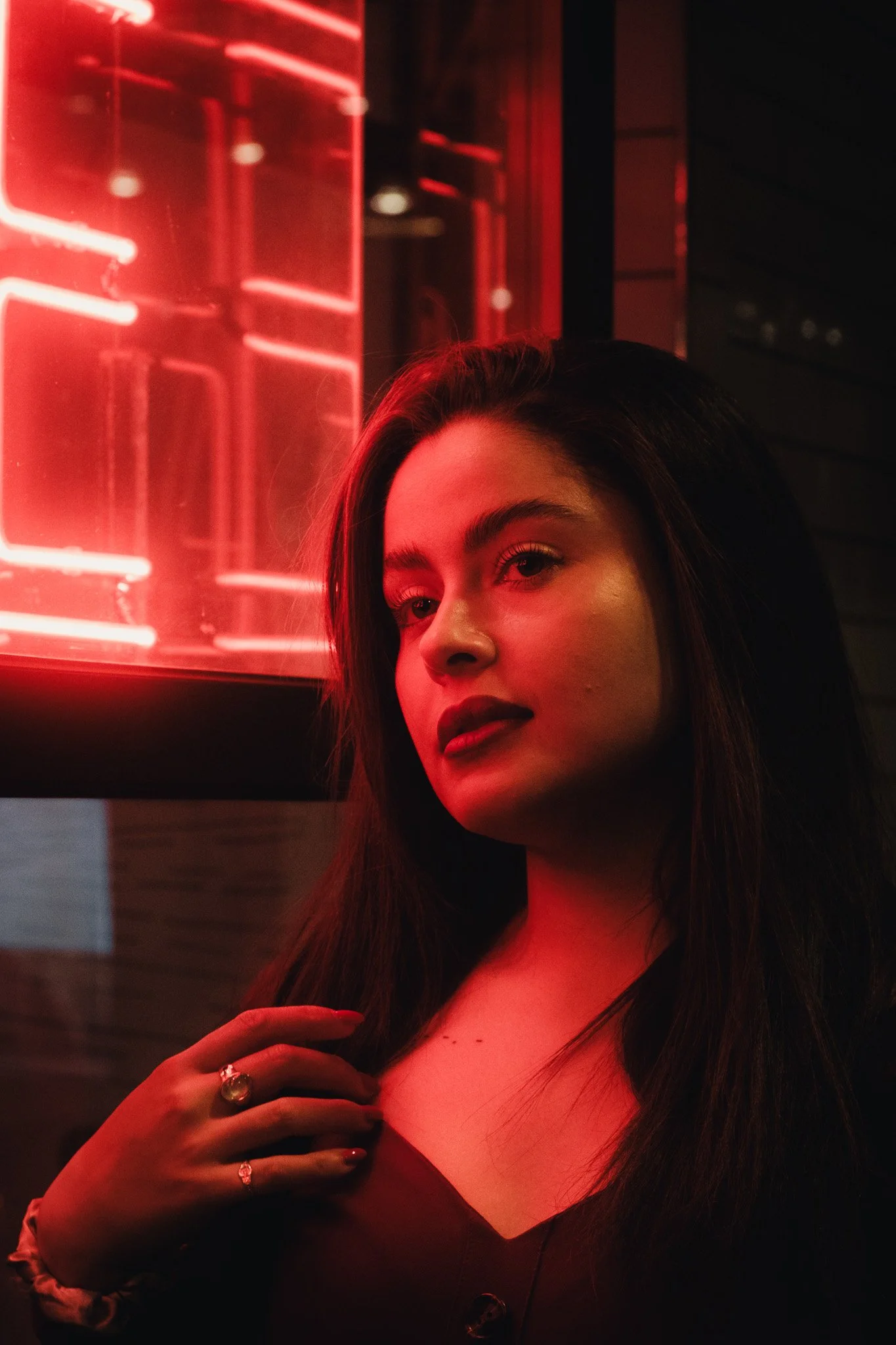 A young woman with dark hair and makeup, standing in front of a red neon light, wearing a dark top, with one hand resting on her chest, adorned with rings and nail polish.