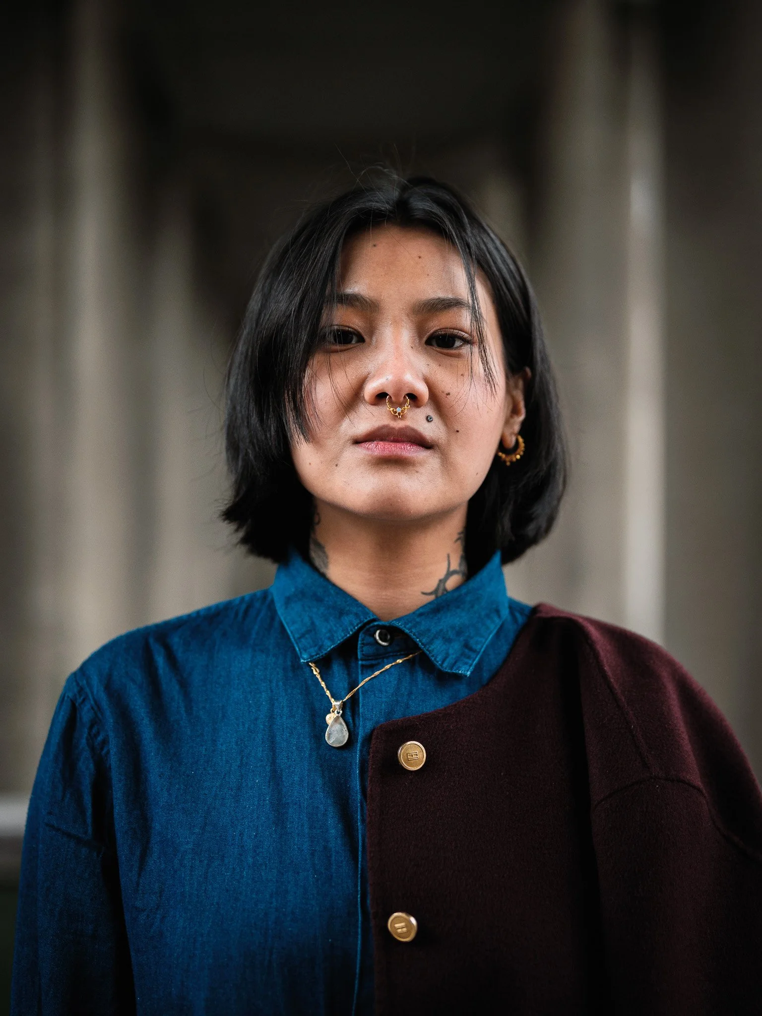 Close-up of a young woman with black hair, septum piercing, gold earrings, wearing a two-toned shirt and a necklace, standing in front of blurred concrete columns.