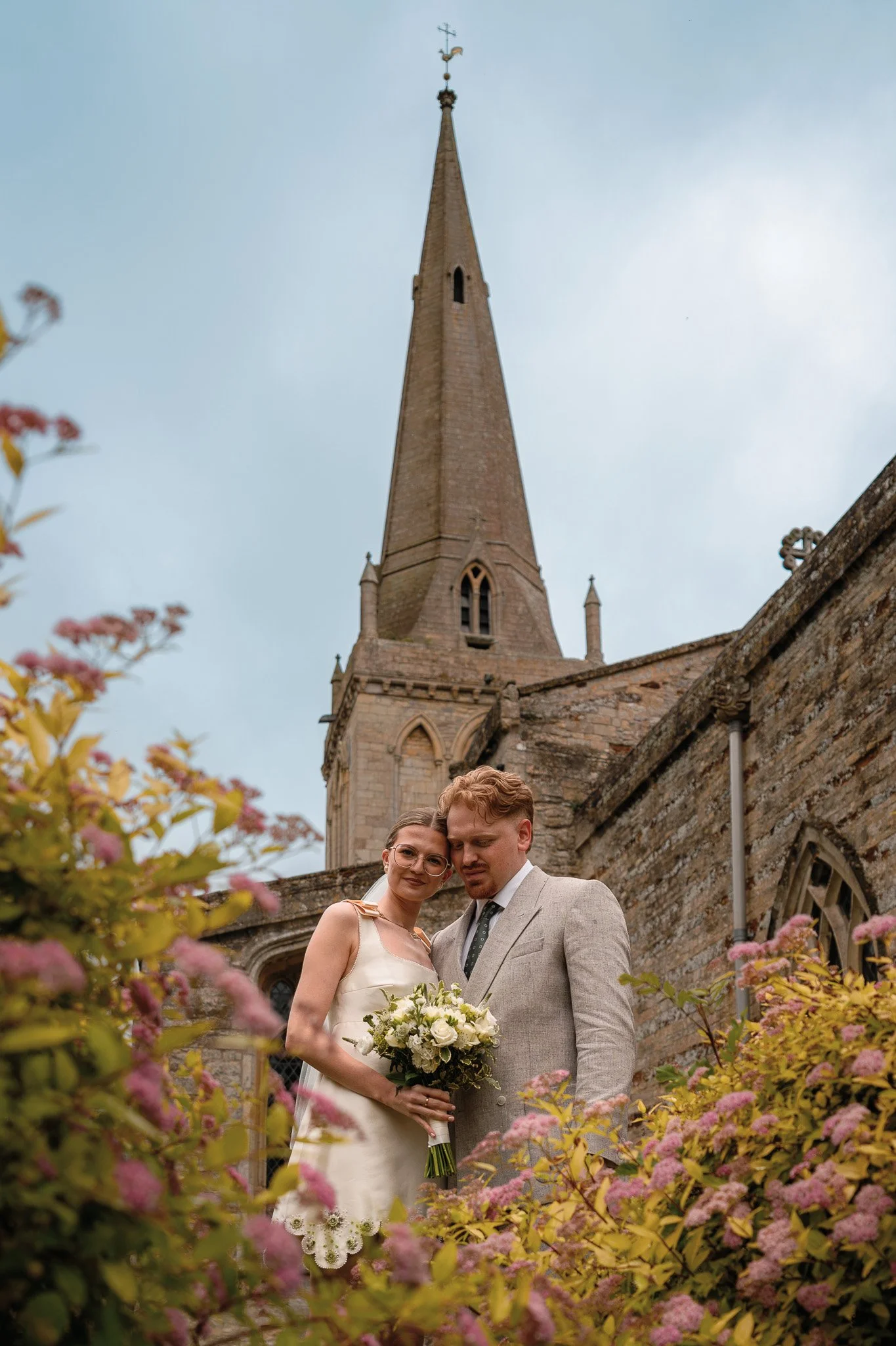 A bride and groom stand closely together in front of an old church, surrounded by pink flowering plants, during a wedding celebration.