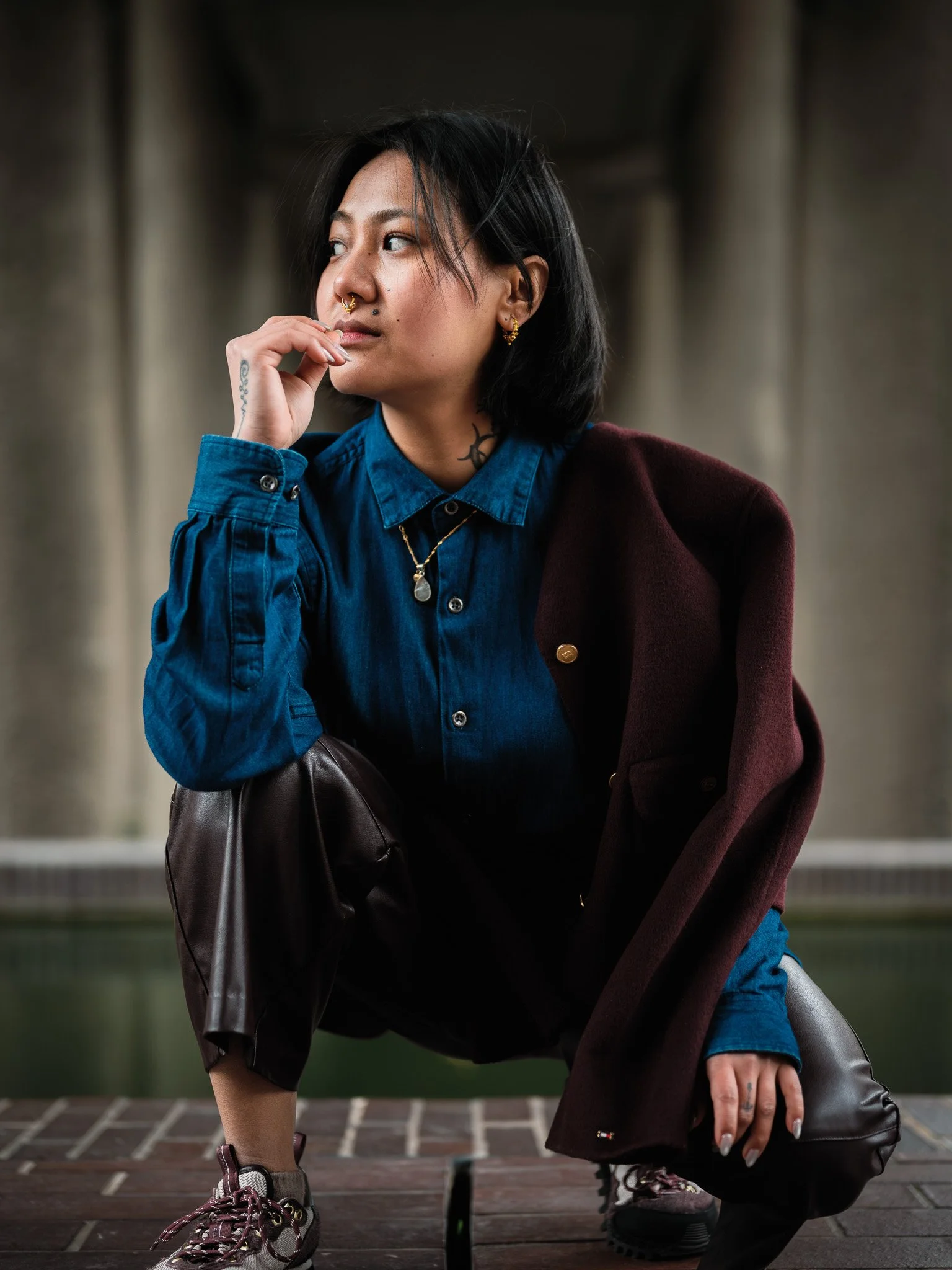A person with black hair, face tattoos, nose piercings, wearing a blue denim shirt layered under a burgundy coat, is squatting on a brick pavement, holding a brown leather bag, with a blurred urban background.
