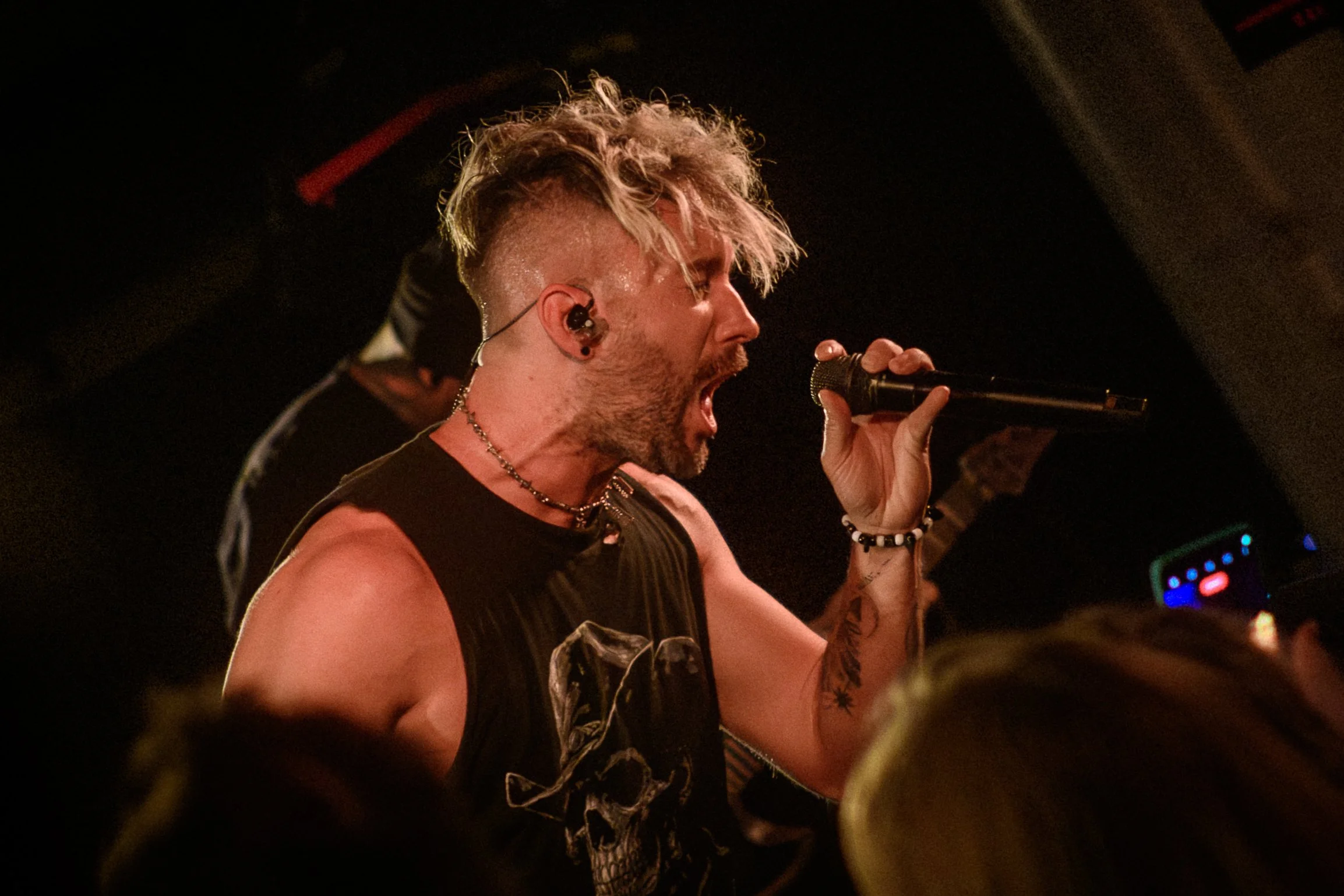 A male singer with bleached blonde hair singing into a microphone on stage, wearing a sleeveless shirt and accessories.