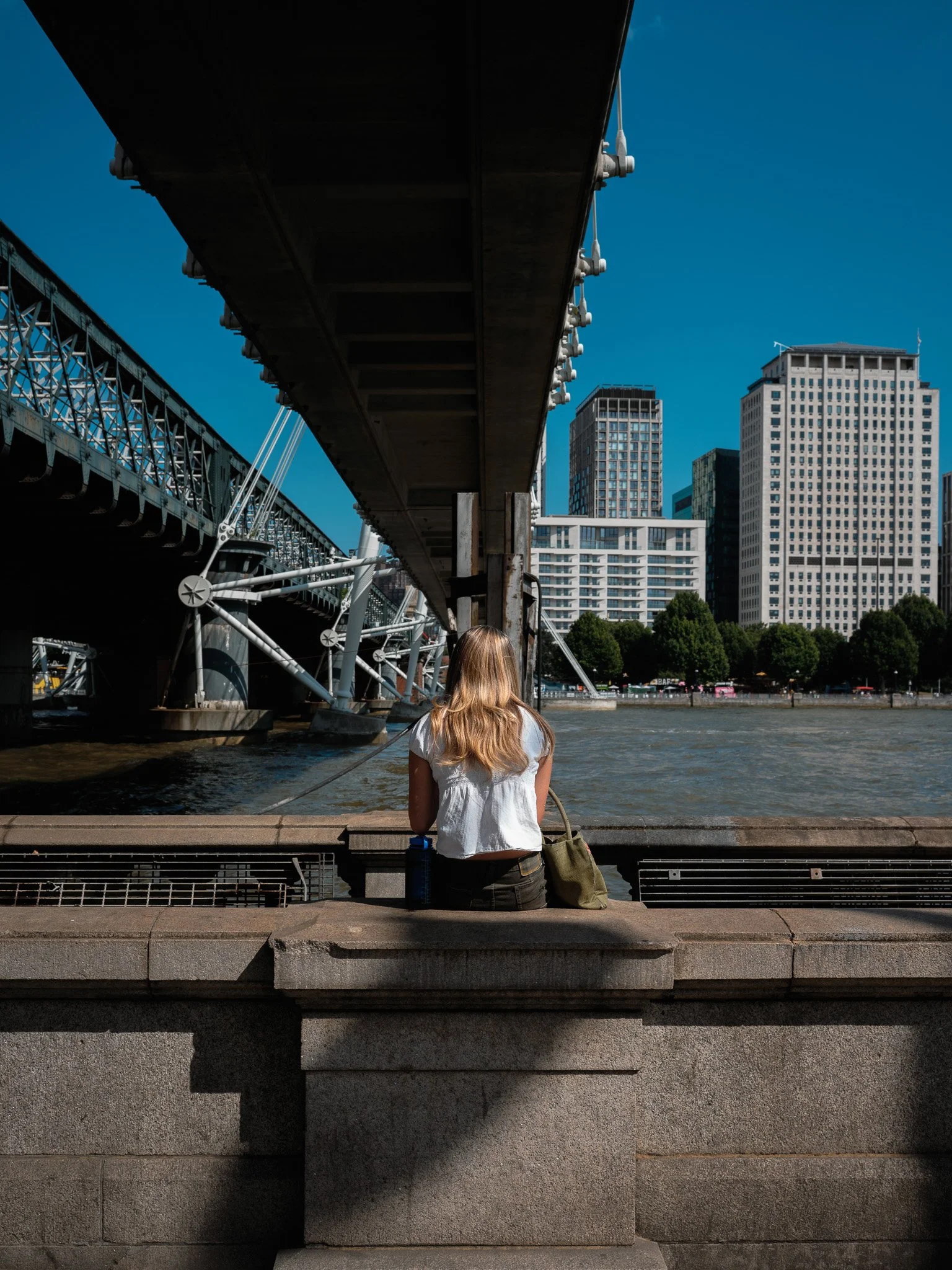 A woman with long blonde hair wearing a white top sitting on a stone bench by a river, facing away, under a bridge in an urban cityscape.