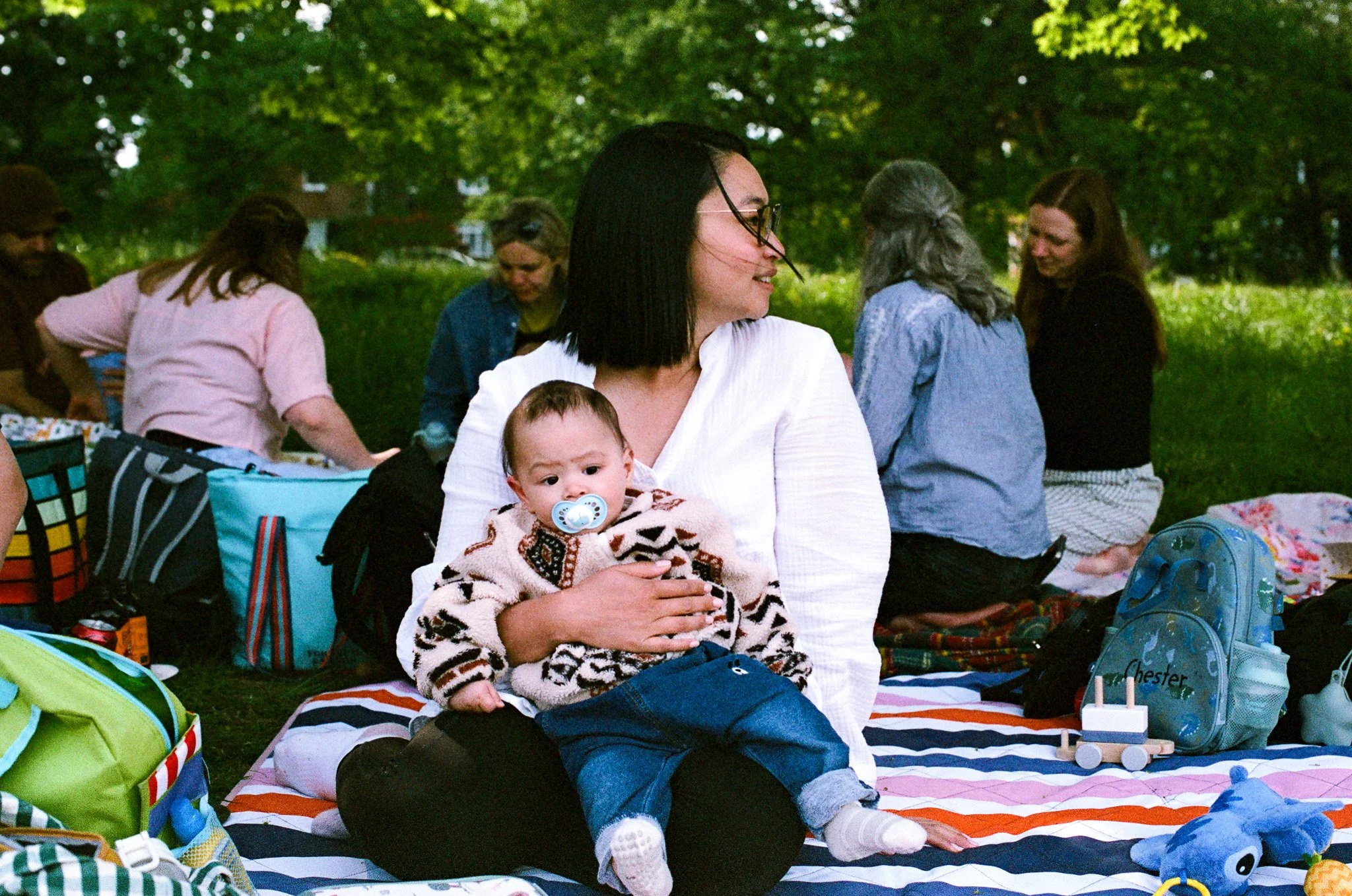 A group of people, mostly women, gathered on a grassy area under trees, having a picnic. A woman with glasses and shoulder-length dark hair holds a baby with a pacifier, sitting on a blanket with various bags and toys around.