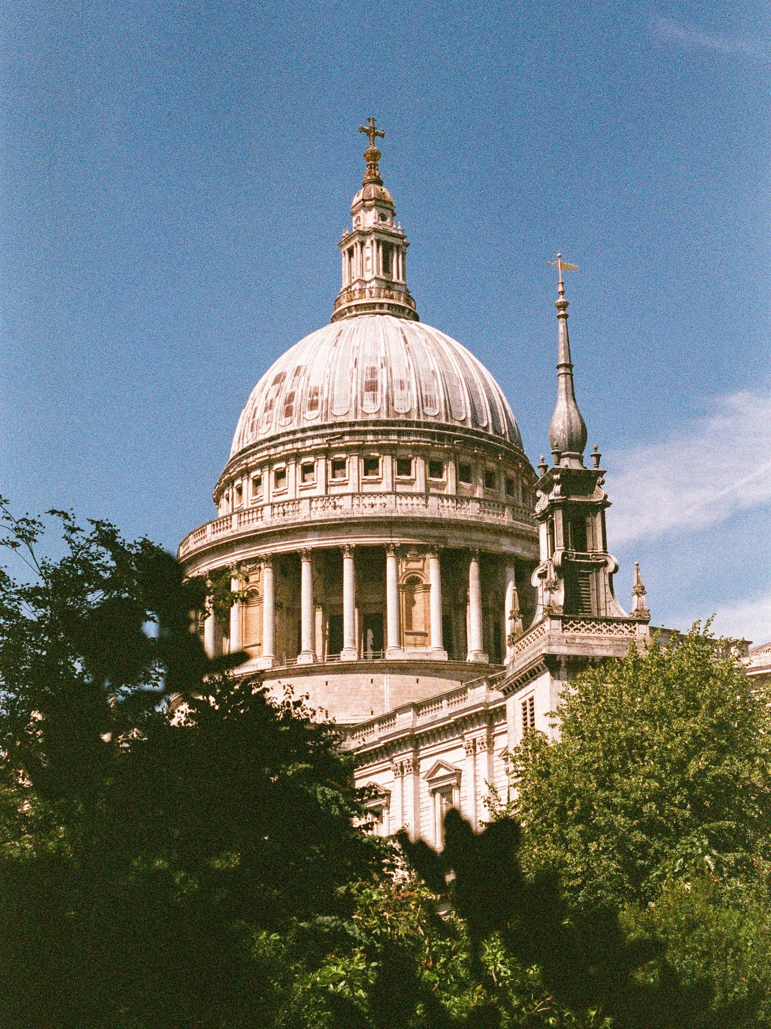 The image shows the dome of St. Paul’s Cathedral in London with a cross at the top, partly obscured by trees in the foreground, under a clear blue sky.