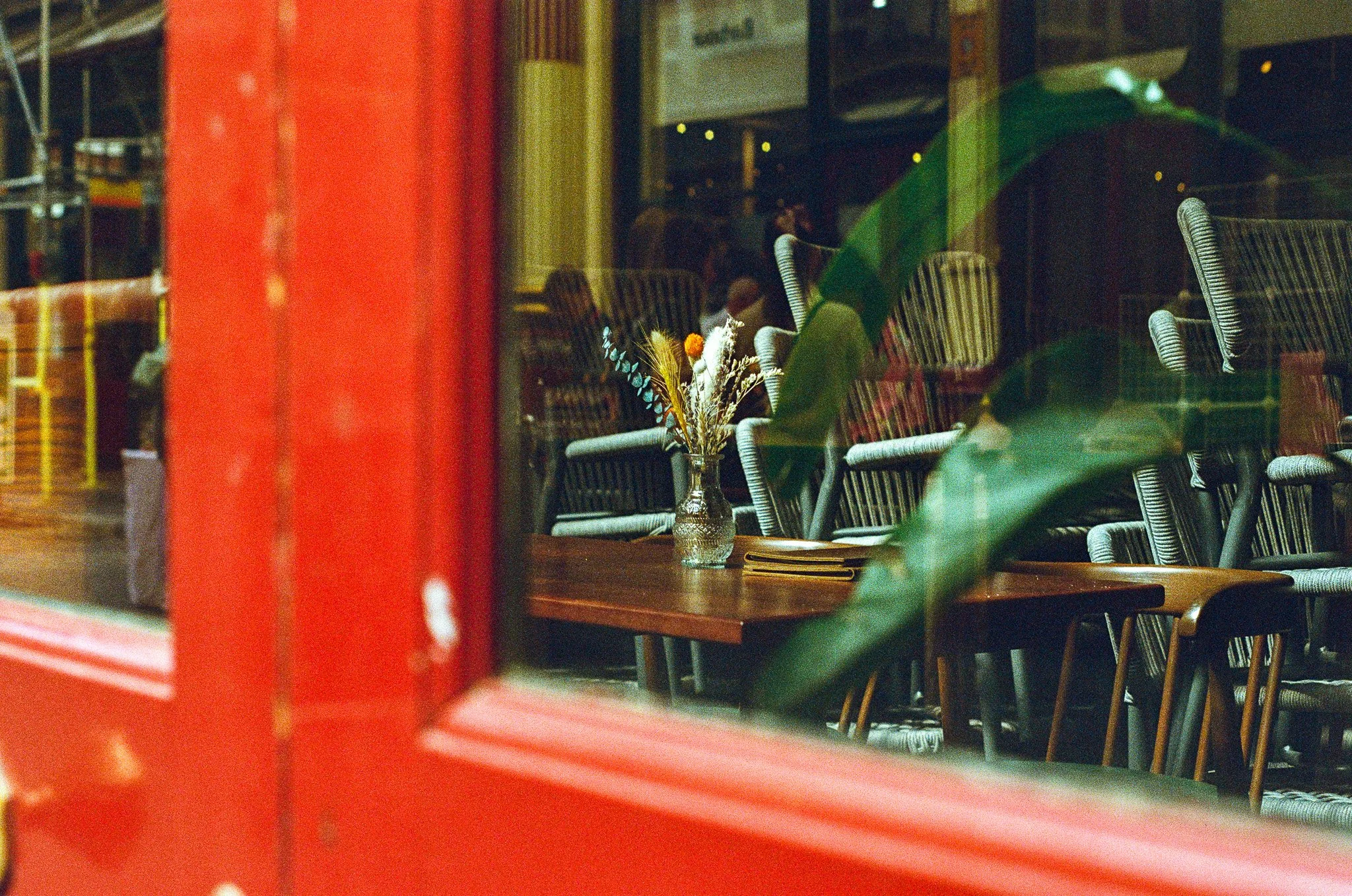 A view through a red window frame showing tables and chairs with a flower vase inside a restaurant at night.