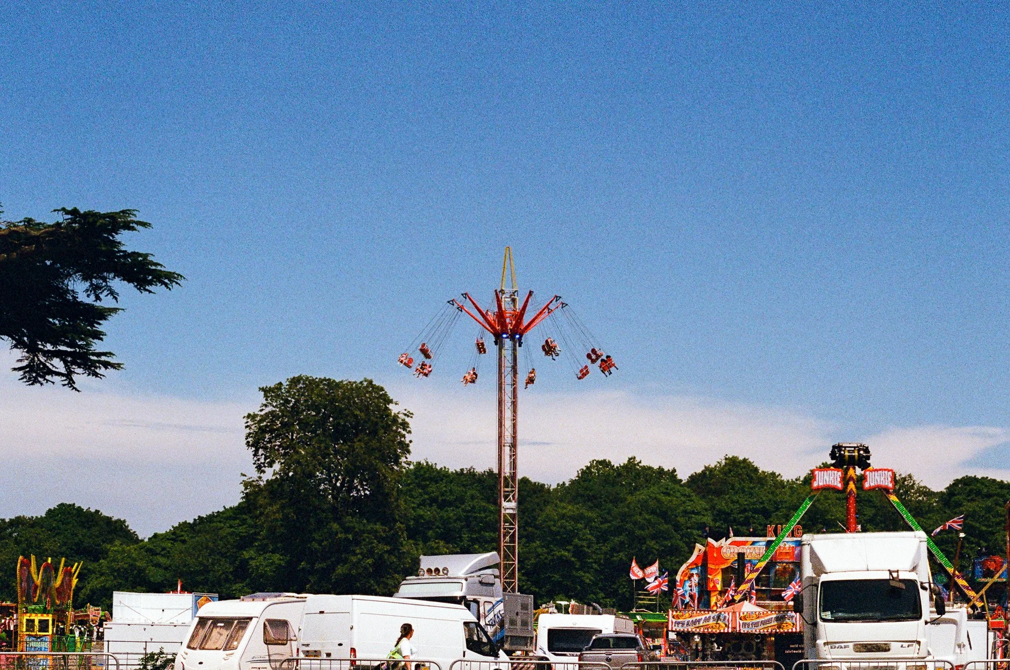 A carnival scene with a large drop tower ride in the center, surrounded by various amusement rides and booths. The sky is clear and blue with trees in the background and several vehicles and people in the foreground.
