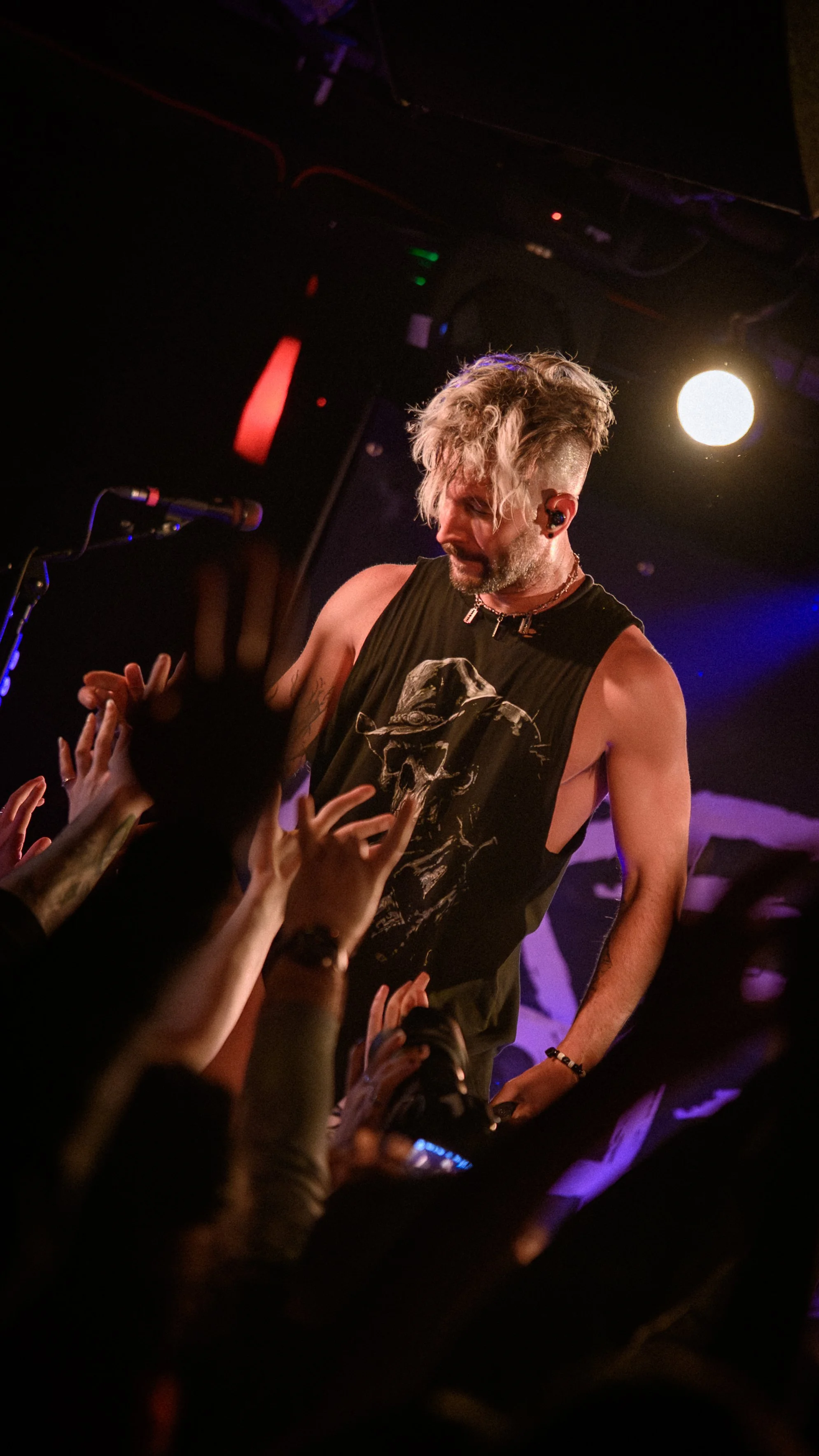 A male performer with bleached, curly hair on stage, reaching out to an audience with hands extended upward, wearing a sleeveless black shirt featuring a skull graphic.