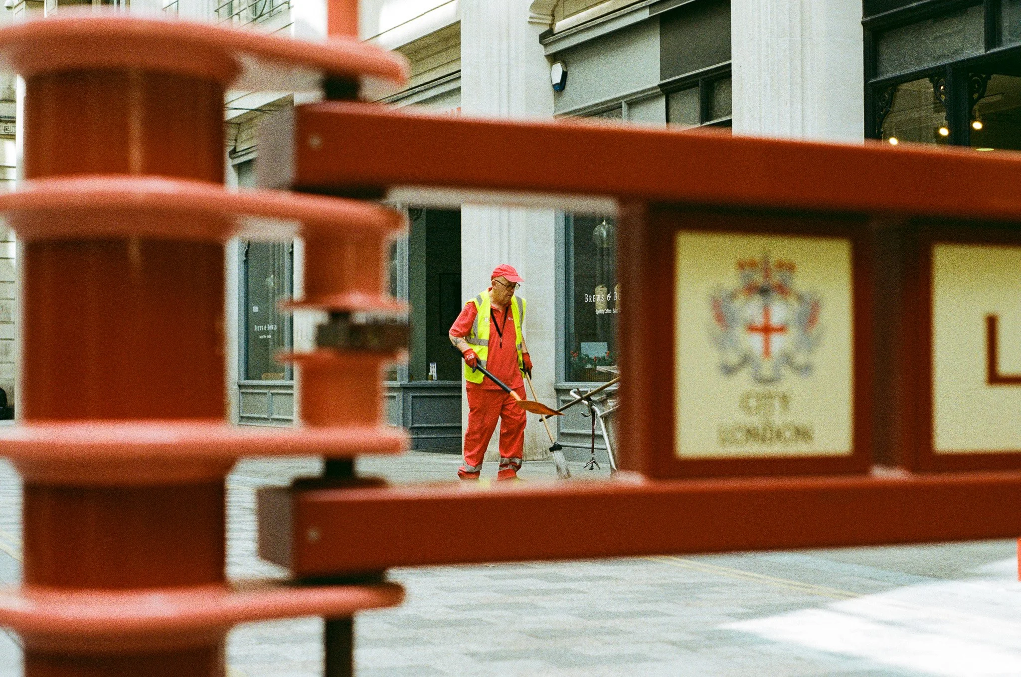 A city street scene seen through a red railing, with a man in a bright yellow safety vest and red clothing sweeping the sidewalk.