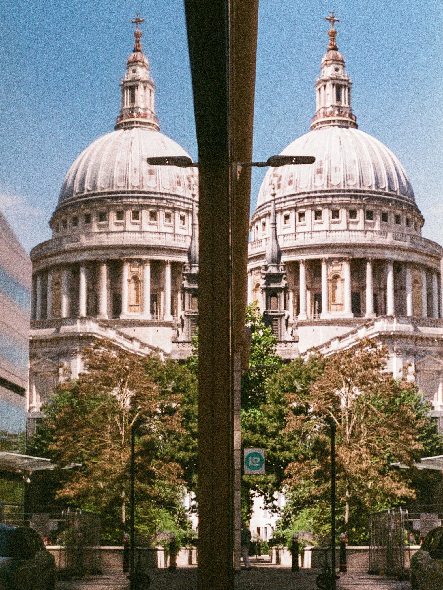 A reflection of a historic domed building with a cross at the top, seen in a mirrored glass surface in an urban setting with trees, cars, and streetlights.