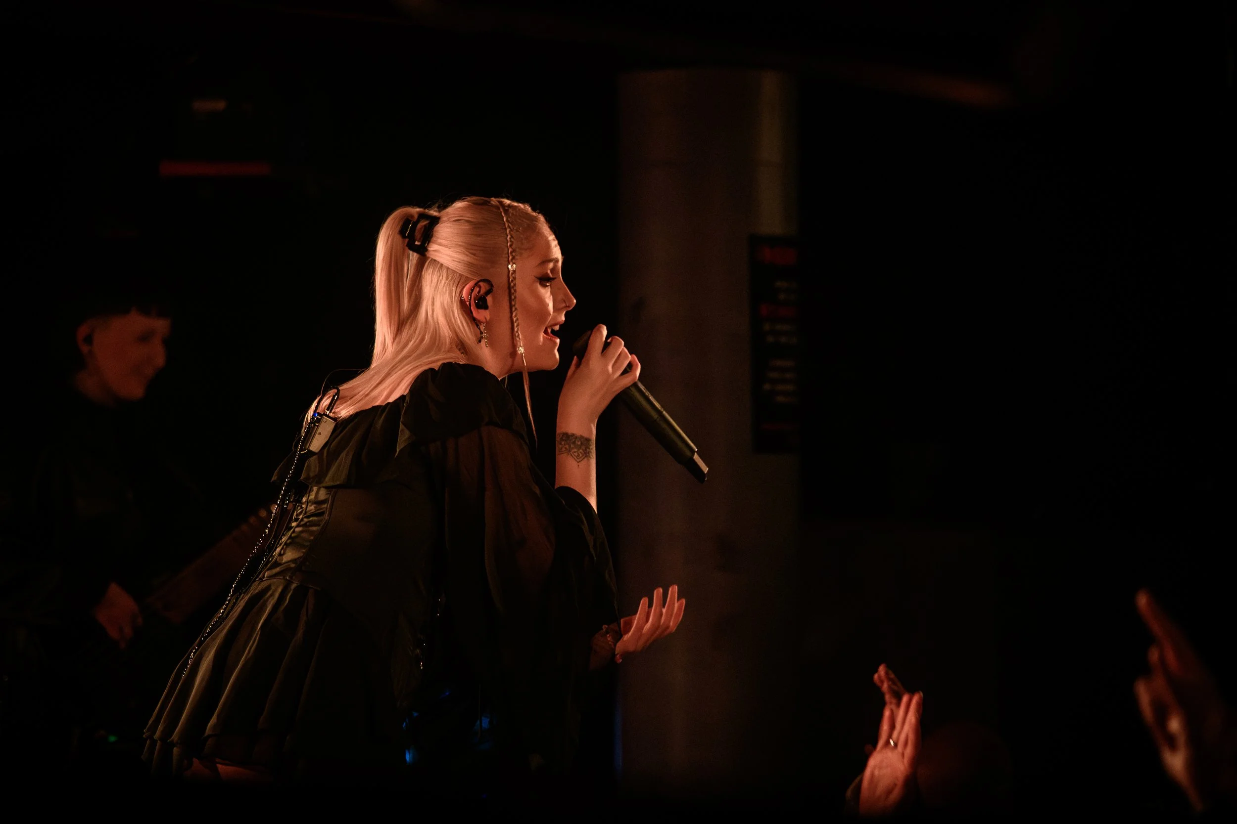A woman singing into a microphone with headphones on, wearing a black jacket, performing on stage. Audience members are raising their hands in front of her.
