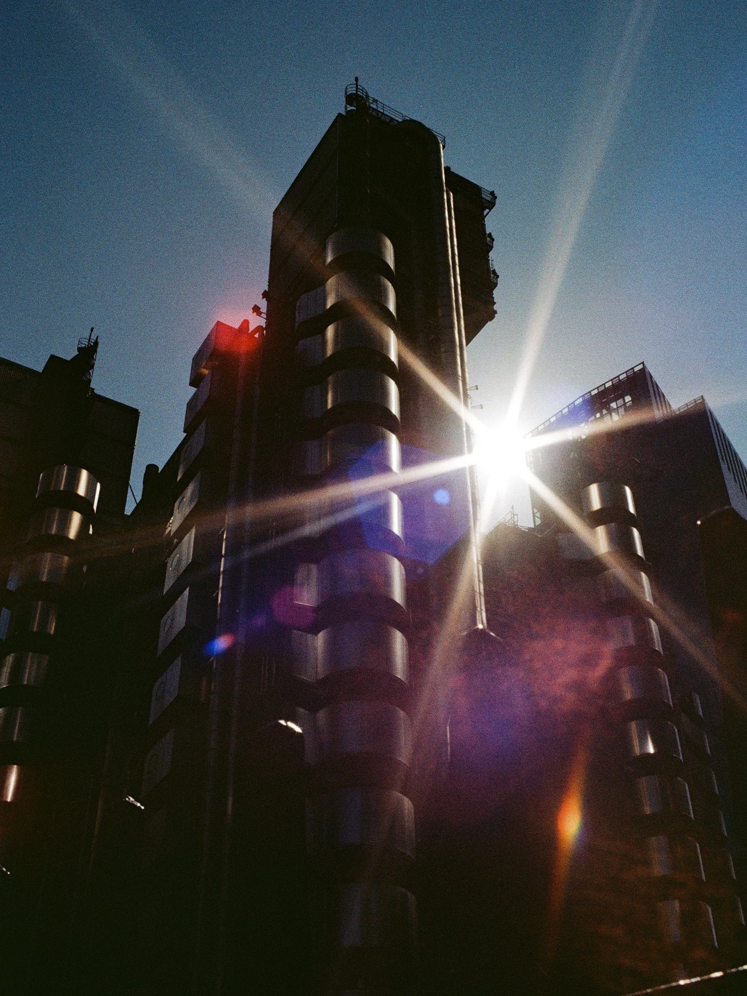 A tall, dark skyscraper with rounded metallic structures along its side, with the sun shining behind it, creating lens flare and casting shadows on the building.