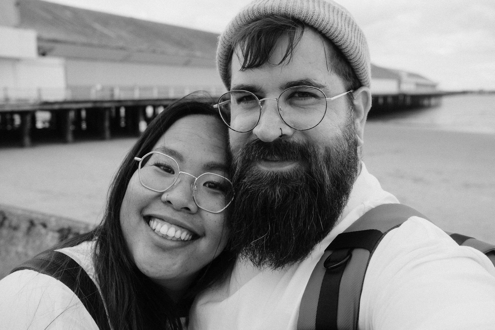 A black and white photo of a smiling woman and a bearded man wearing glasses and a beanie, taking a selfie outdoors near a bridge.