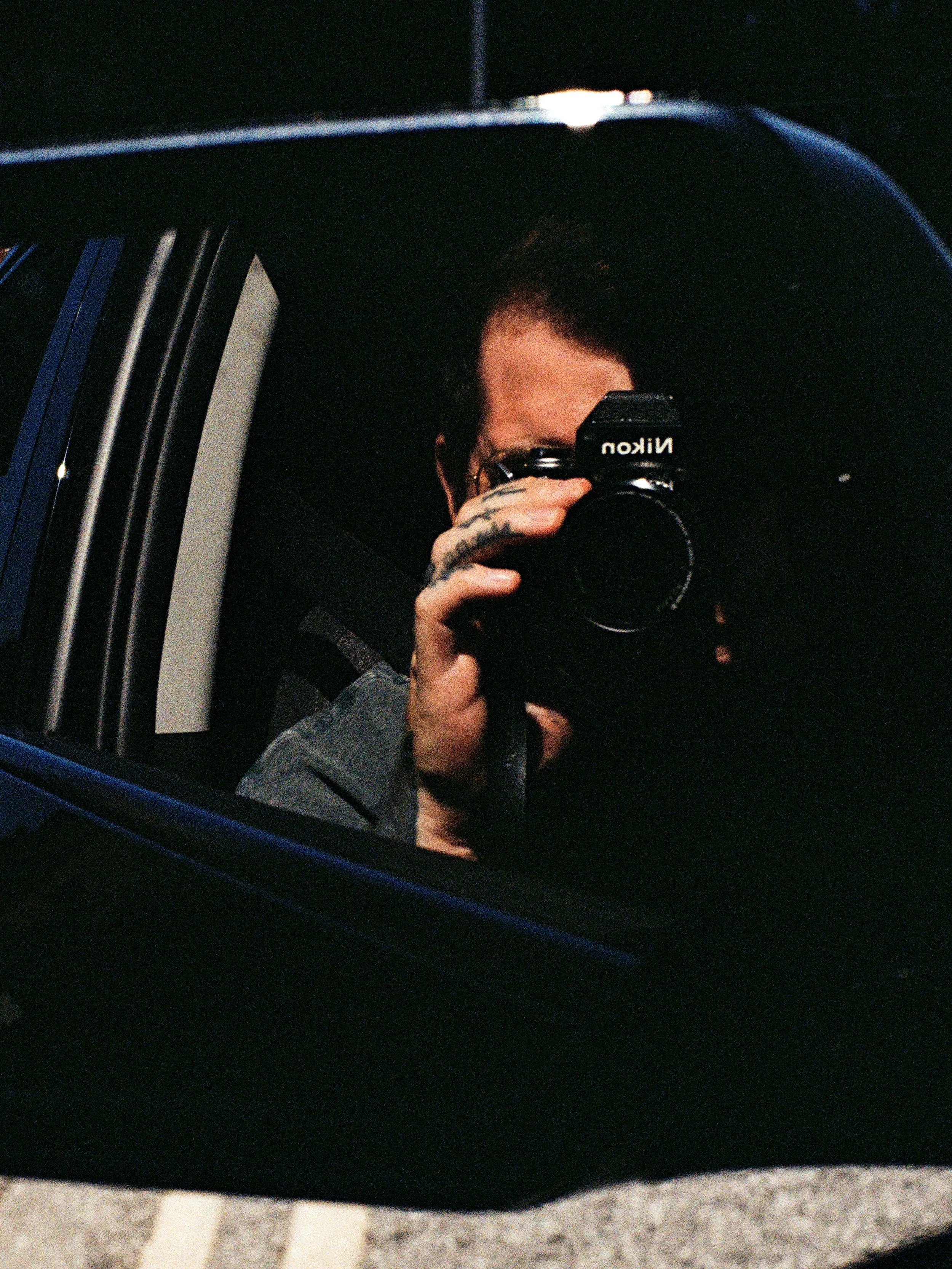 A person taking a photograph of their reflection in a car's side mirror. The person has tattoos on their hand and is holding a Nikon camera. The background appears dark, possibly at night.