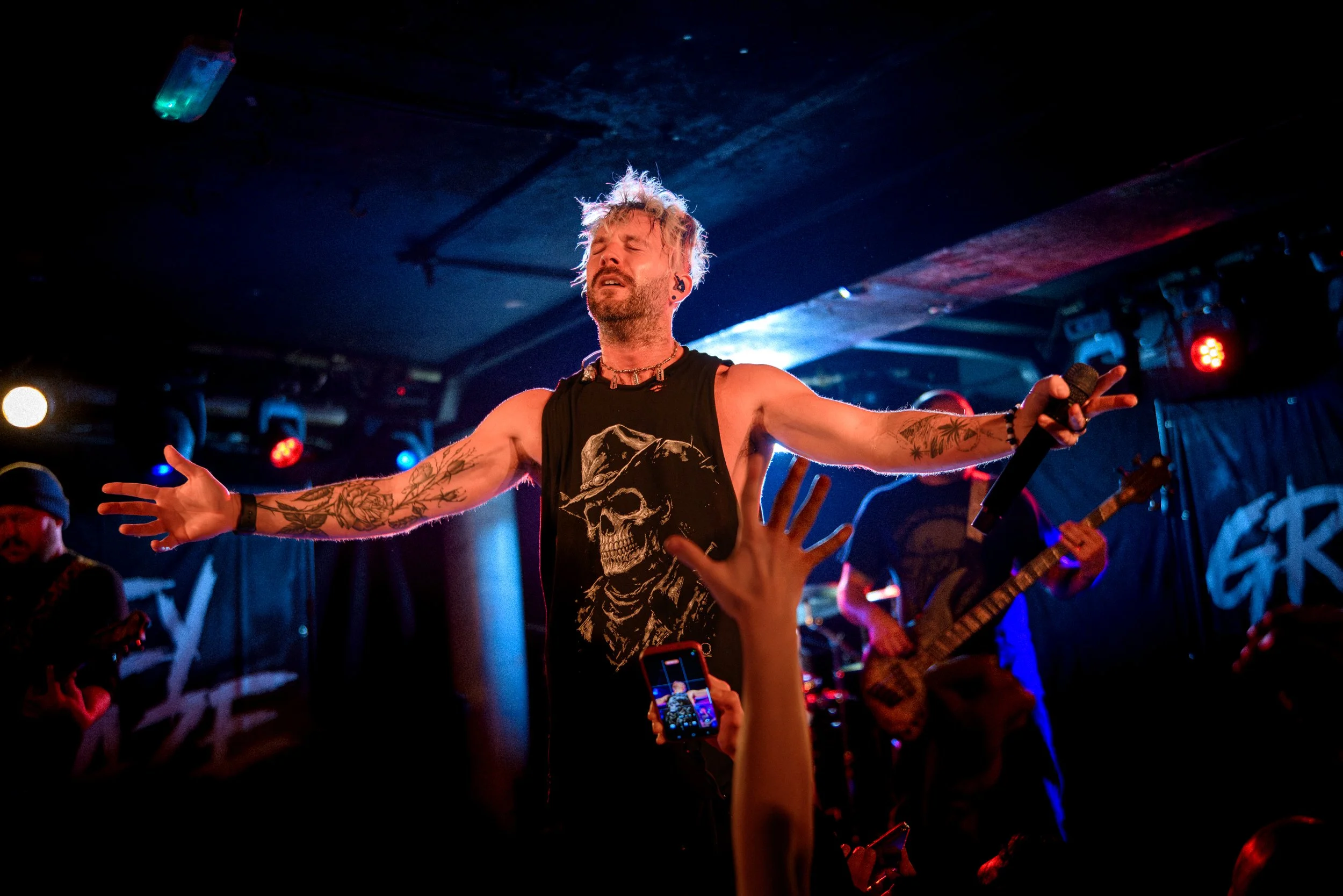 A male singer with tattooed arms, wearing a sleeveless shirt with a skull graphic, performs passionately on stage with eyes closed. He holds a microphone with his right hand, and his left arm is extended outward. The stage is dimly lit with colorful 