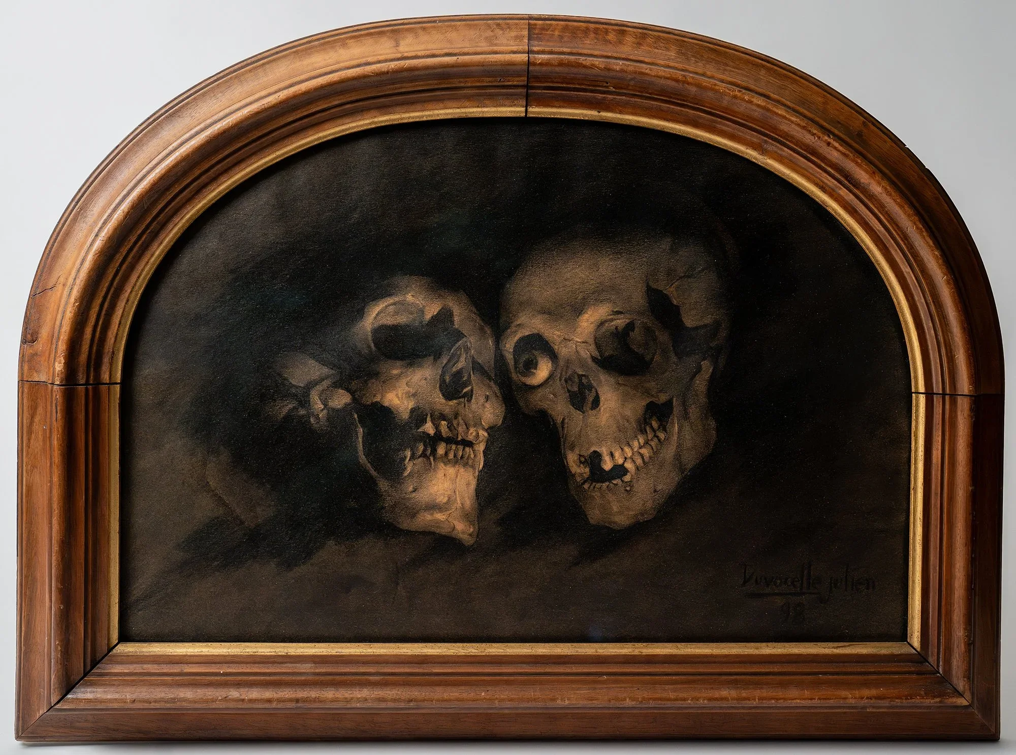 Oil painting of two human skulls against a dark background in an arched wooden frame