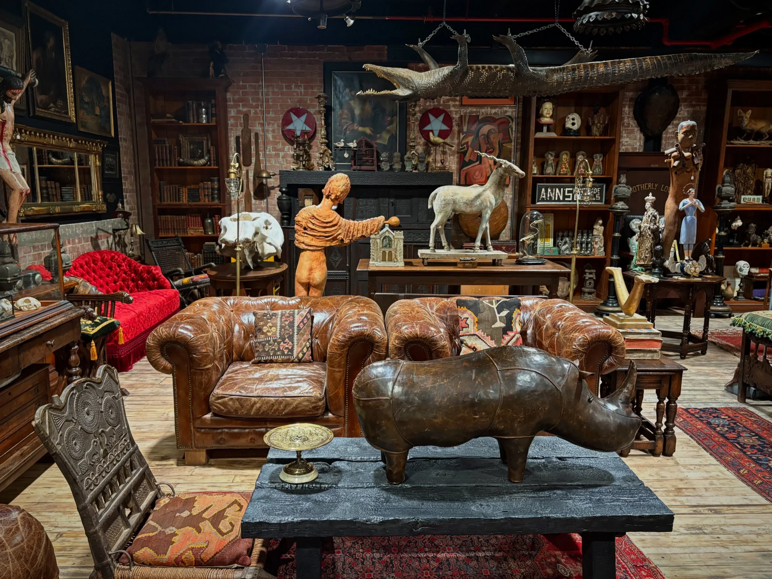 An eclectic collection of vintage and antique items in a room with an exposed brick wall, including a large hanging crocodile, statues, paintings, books, and various decorative objects.