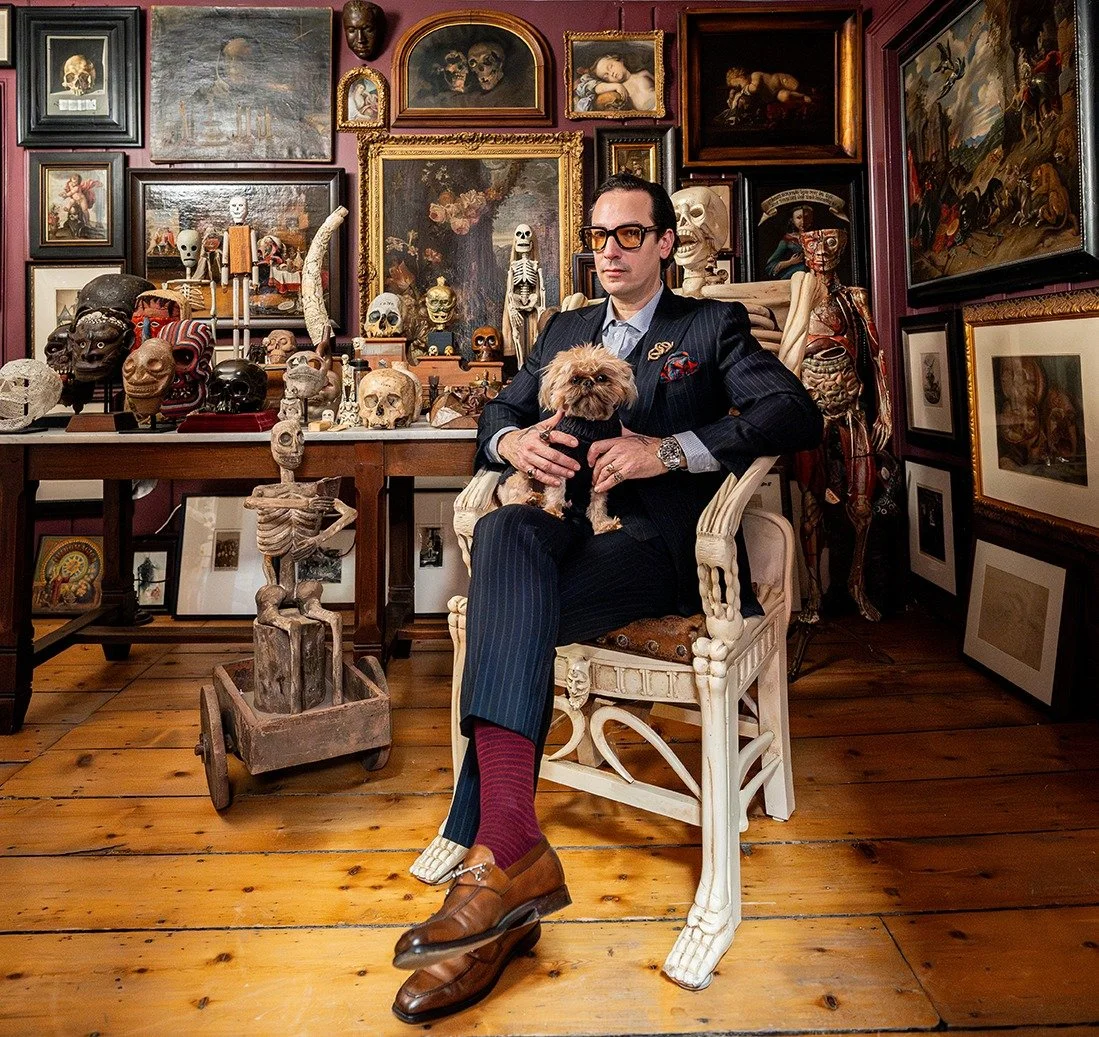 Ryan Matthew Cohn in a pinstripe suit, seated with his dog among skulls, paintings and curiosities in his collection
