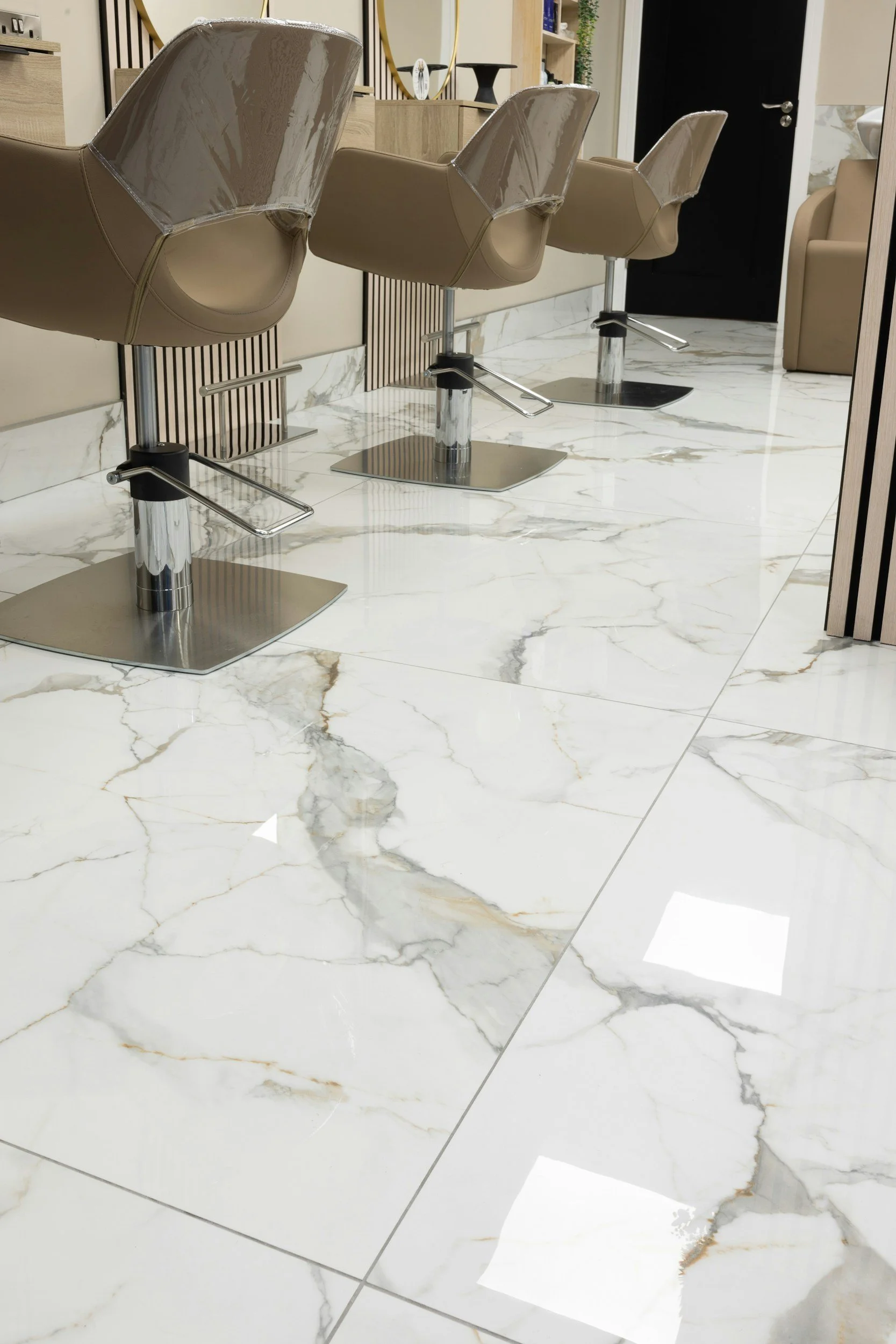Three salon chairs with beige upholstery and chrome bases in a salon with white marble floor tiles and decorative wall panels.