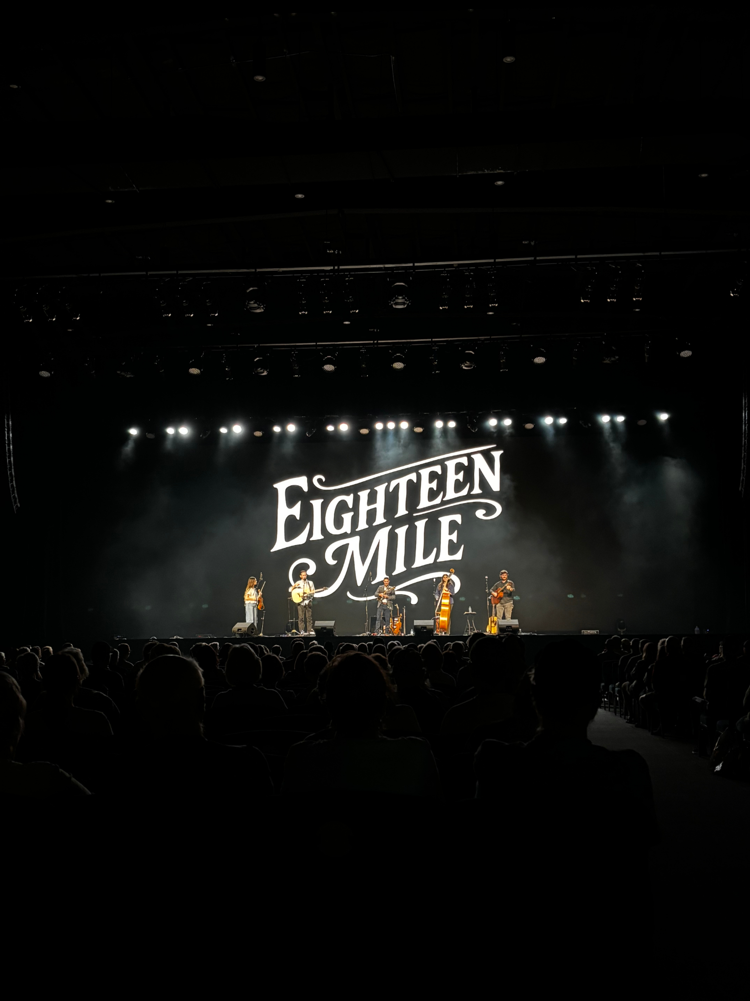 A band of five musicians performing on stage with the backdrop displaying the words 'FIGHTEEN MILE'. The audience is watching from in front of the stage in a darkened concert hall.