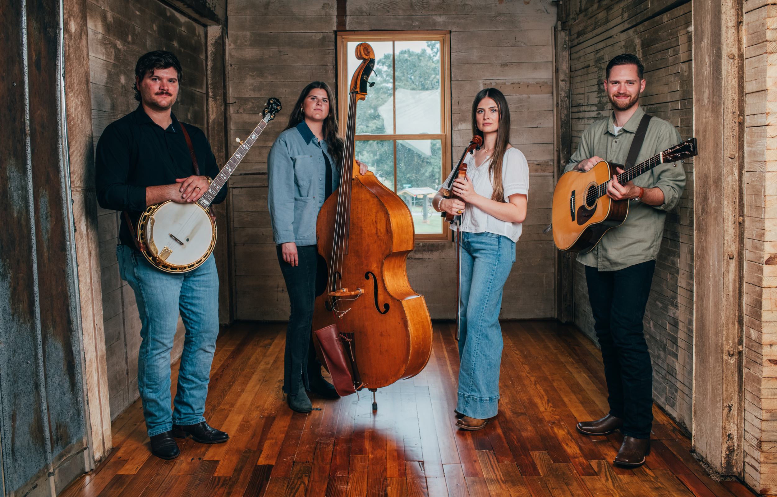 A group of four musicians standing in a rustic wooden room, each holding a different musical instrument, including a banjo, double bass, violin, and acoustic guitar.