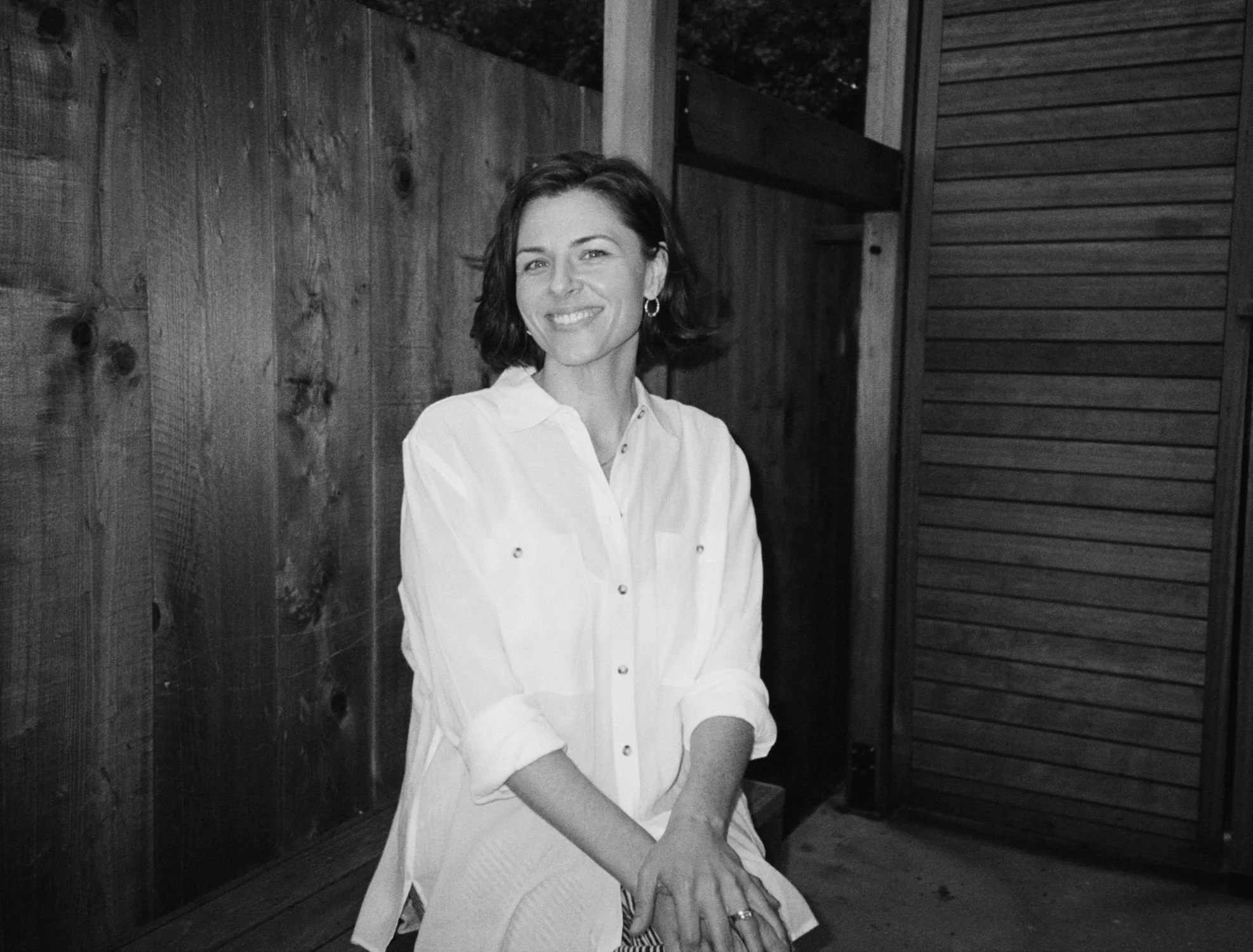 A woman sitting outdoors on a wooden bench, smiling, wearing a white shirt, with wooden fencing and a building with horizontal siding in the background.