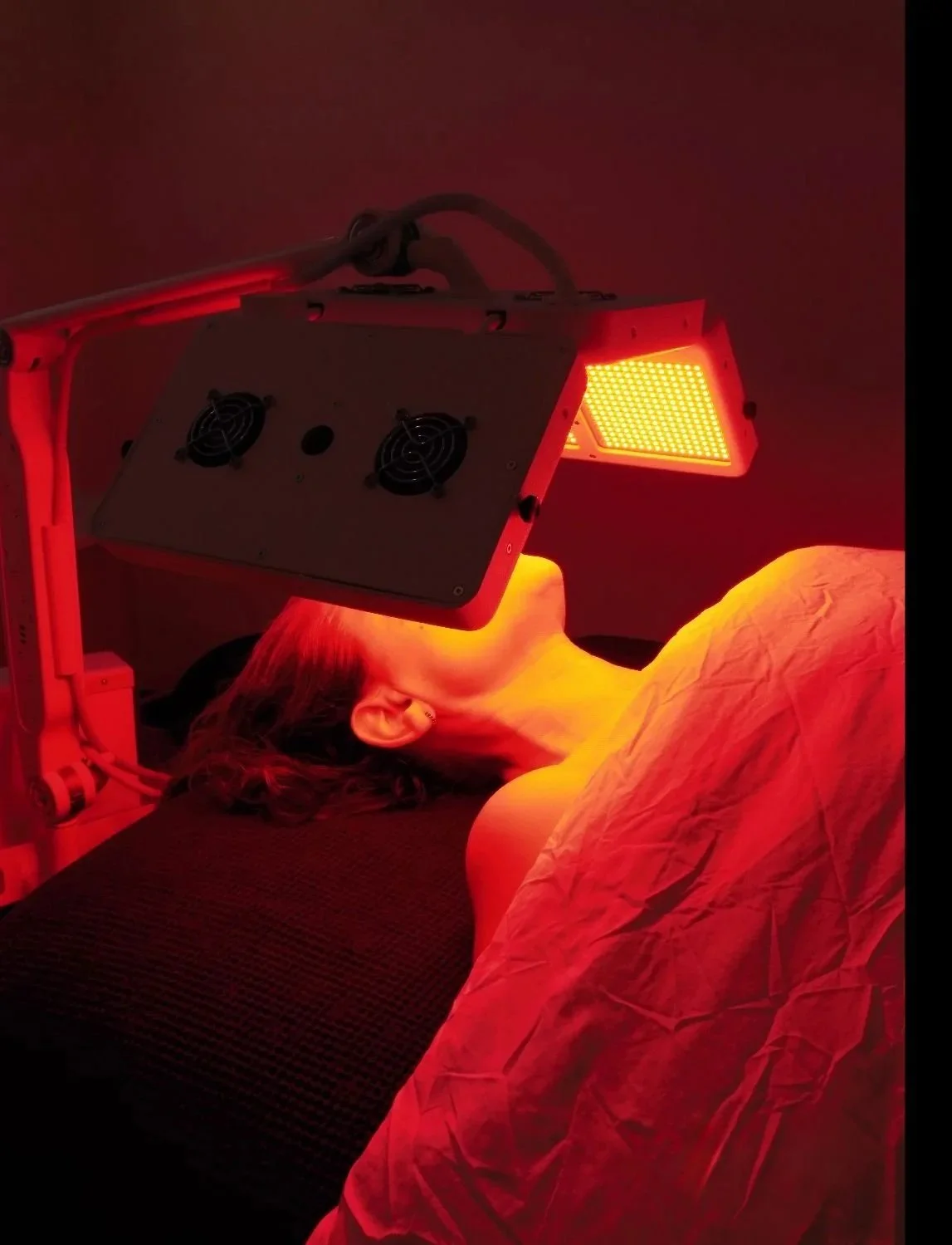 Person lying on a bed undergoing red light therapy, with a red LED light panel above their face.