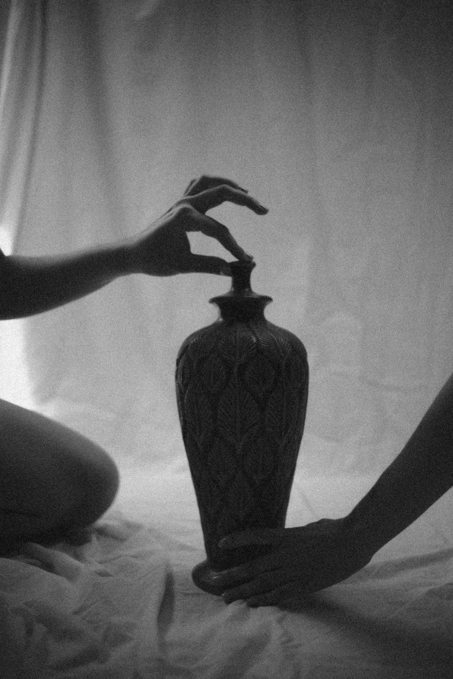Black and white photo of a hand balancing on an ornate vase with a paper roll nearby, against a plain background.