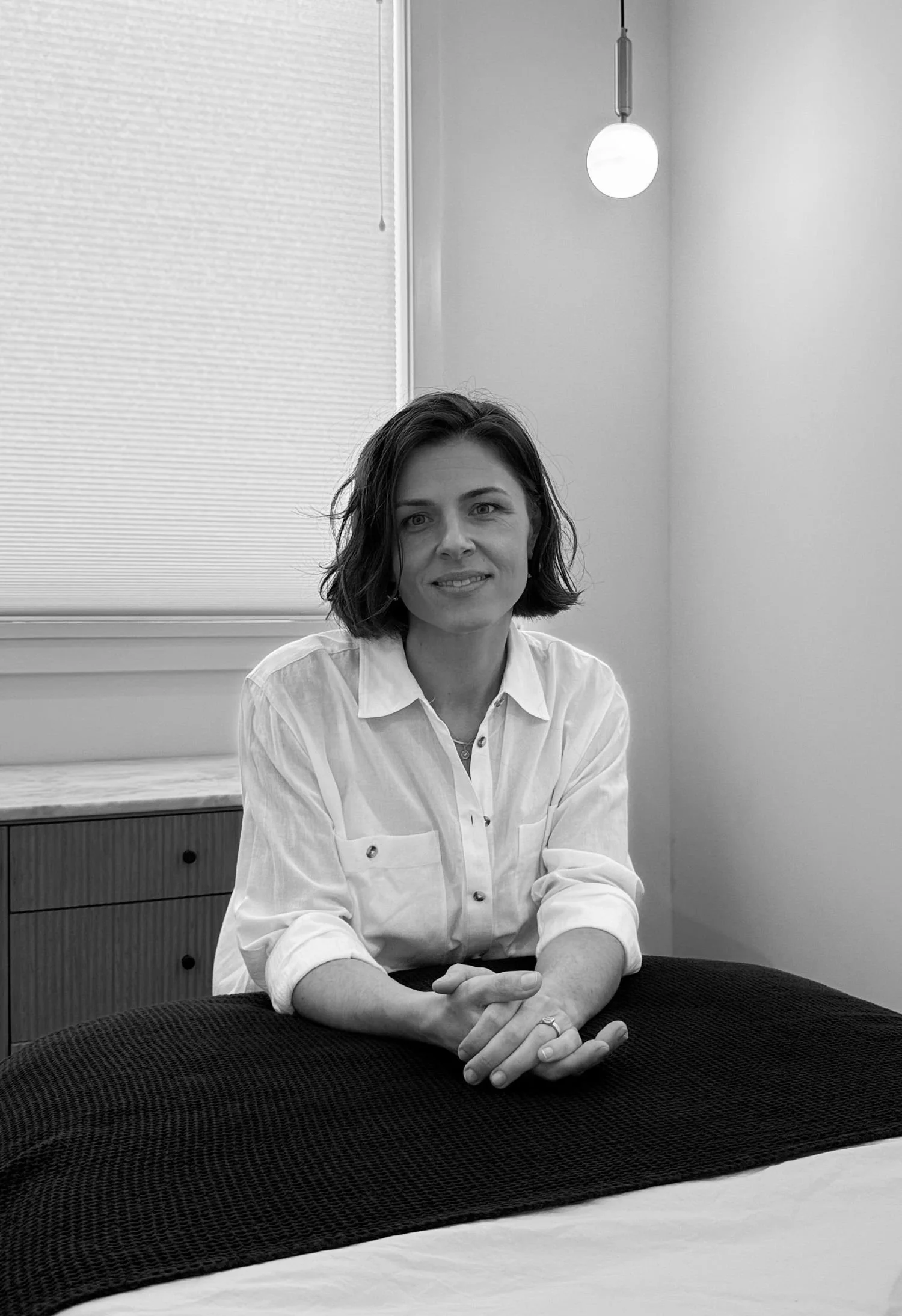A woman sitting at a table with hands folded, wearing a white button-up shirt in a room with a window and ceiling light. The photo is in black and white.