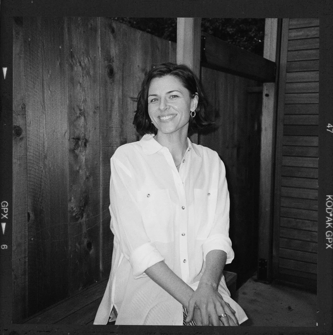 Black and white photo of a woman with short dark hair, wearing a white shirt, smiling and sitting outdoors against a wooden fence.