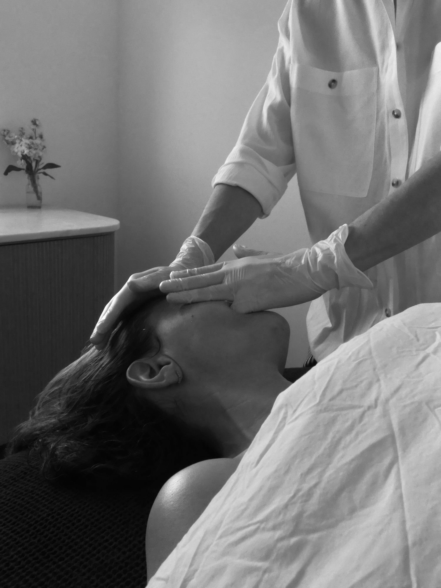 A healthcare professional wearing gloves providing treatment or massage to a patient lying on a bed, with a flower vase on a table in the background.