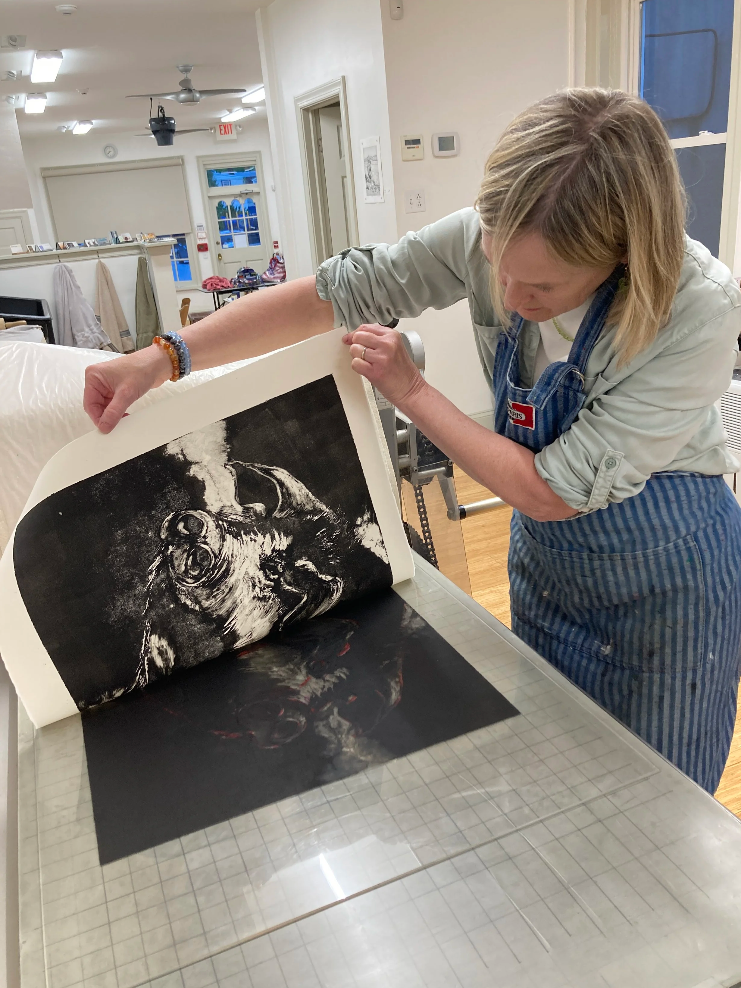 A woman with blonde hair and a striped apron carefully handling a black and white artwork of a pig, at print studio and gallery.