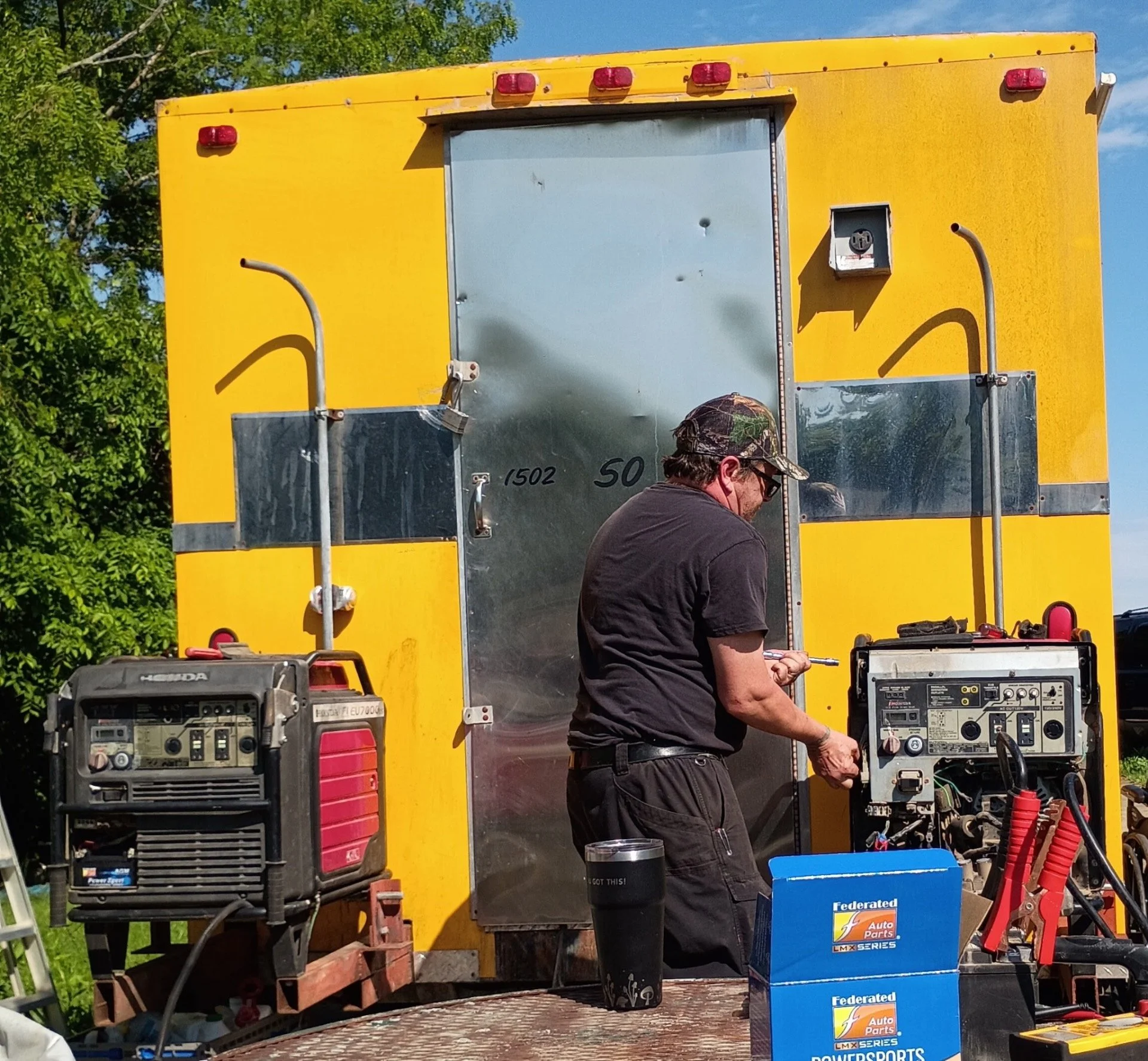 A man in black shirt, glasses, and camouflage cap working on a car engine outside near a yellow food truck or trailer with trees and blue sky in the background.