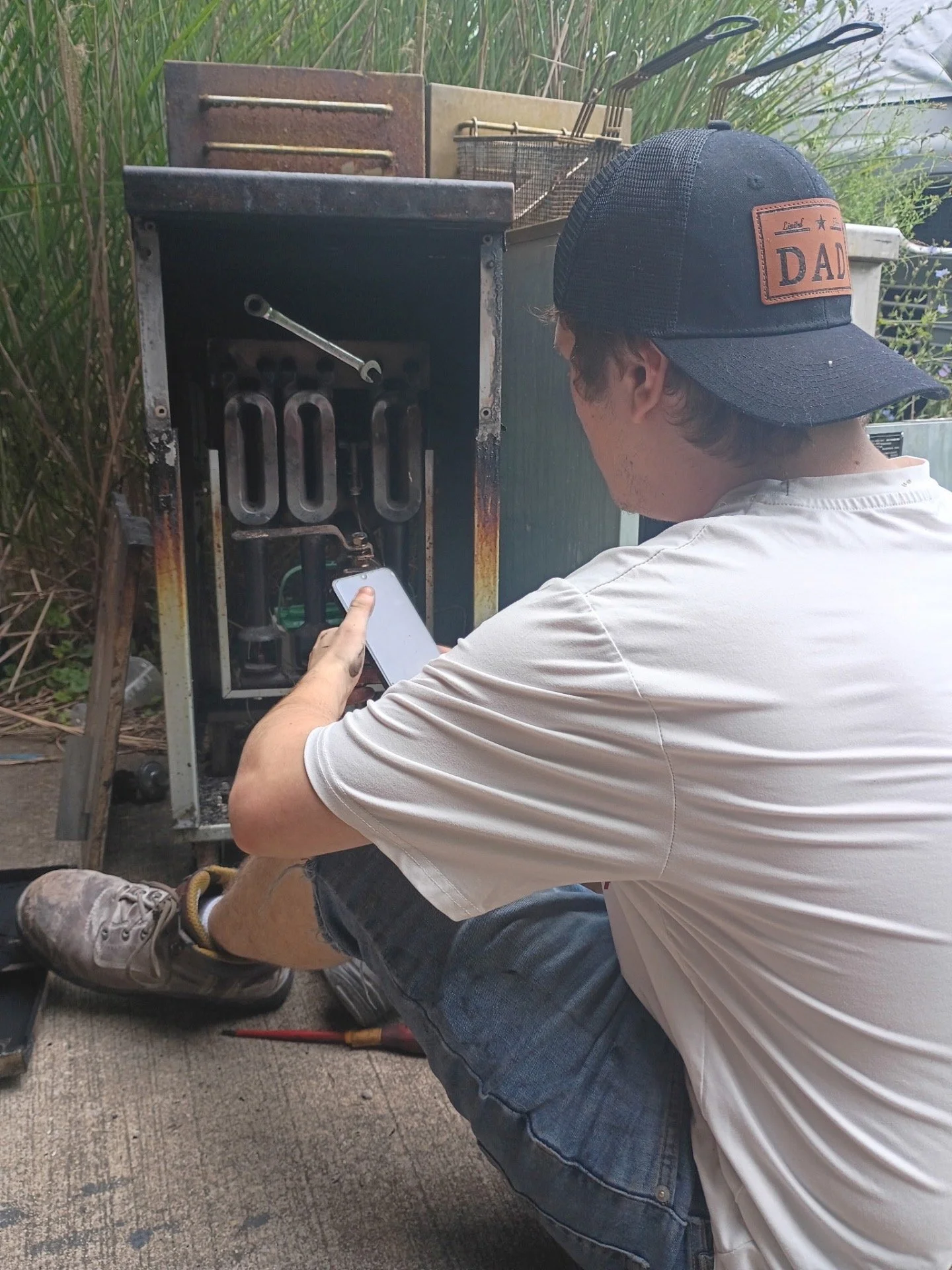 A man in a white t-shirt, black shorts, and gray sneakers working on an outdoor furnace or metal cabinet, wearing a black baseball cap with a leather patch labeled DAD, sitting on the ground with a smartphone in his hand.