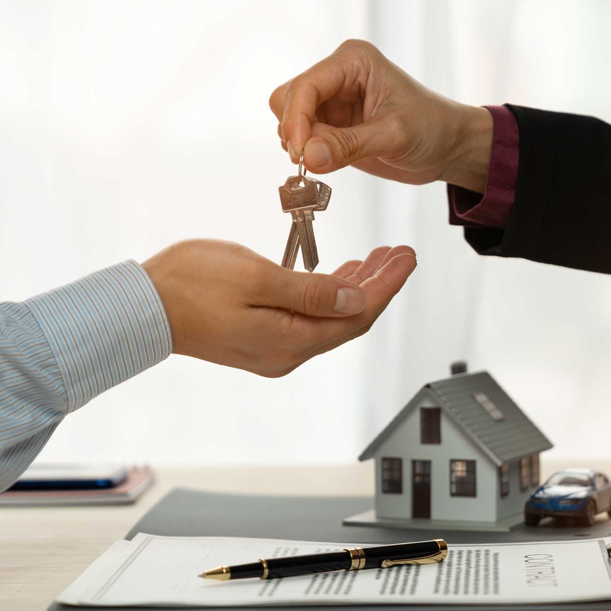 Person handing over a set of house keys to another person in an office setting with a model house and office supplies in the background.