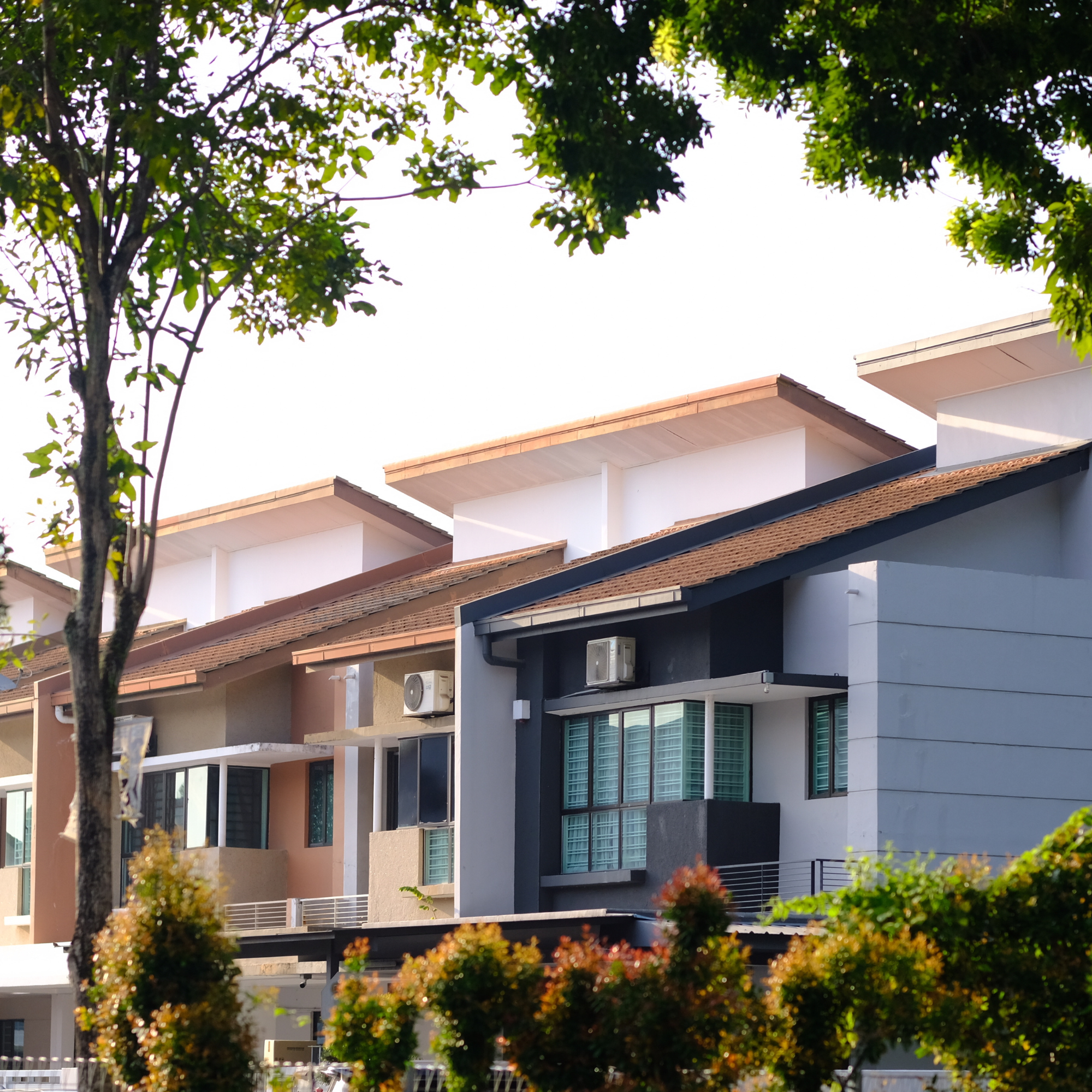 Multi-story residential building with white and dark exterior finishes, multiple large windows with green curtains, situated outdoors with green trees and plants in the foreground.