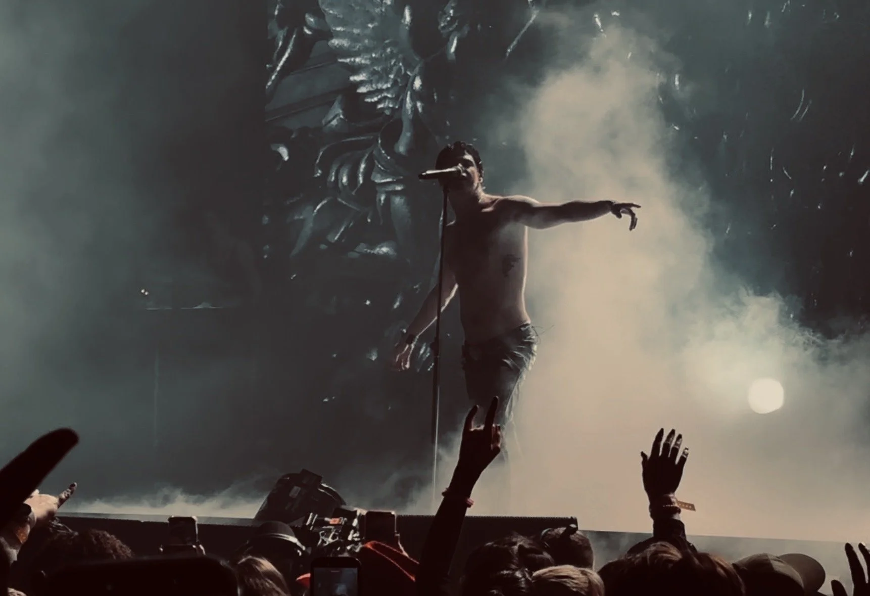 A shirtless male performer singing into a microphone on stage, surrounded by fog and smoke effects, with an audience reaching out towards him.