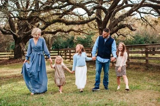 A family of six walking hand-in-hand outdoors in a grassy area with large trees and a wooden fence in the background.