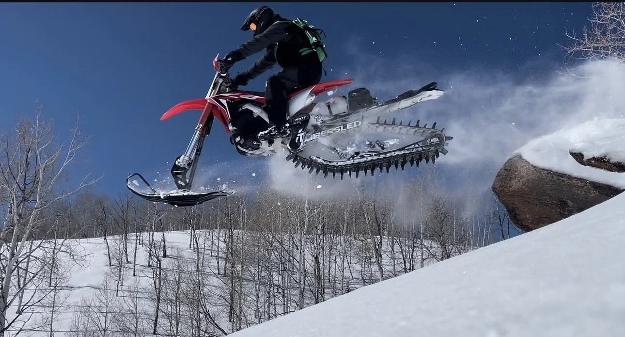 A person in winter gear riding a snowmobile jumps off a snow-covered rock in a snowy landscape with leafless trees and a blue sky.