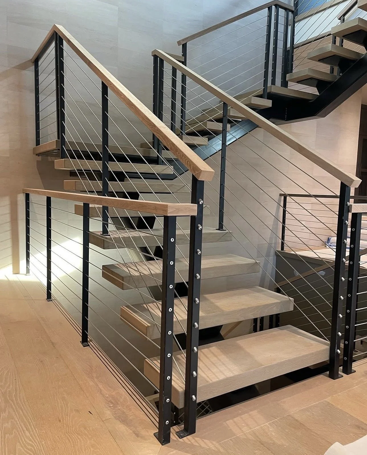 Modern staircase with wooden steps, black metal frame, and cable railing, inside a building with wood flooring and wood-paneled walls.