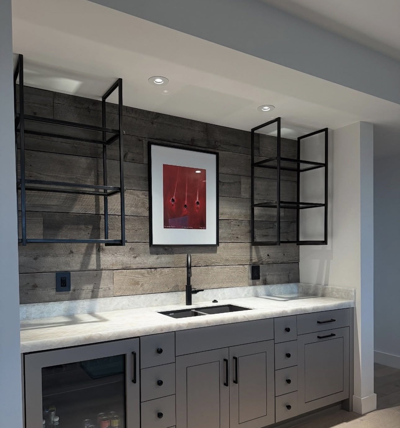 Modern kitchen with gray cabinetry, white marble countertop, black faucet, wood accented wall, open black metal shelves, and a framed red art print.
