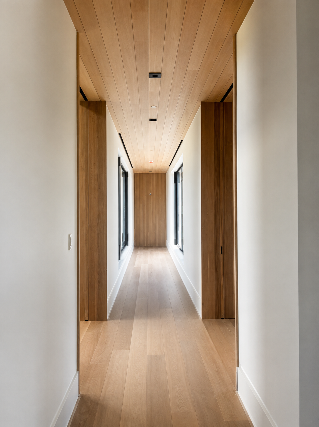 A modern, minimalistic hallway with light wooden flooring, white walls, and wooden ceiling and accents. Large windows on both sides let in natural light.