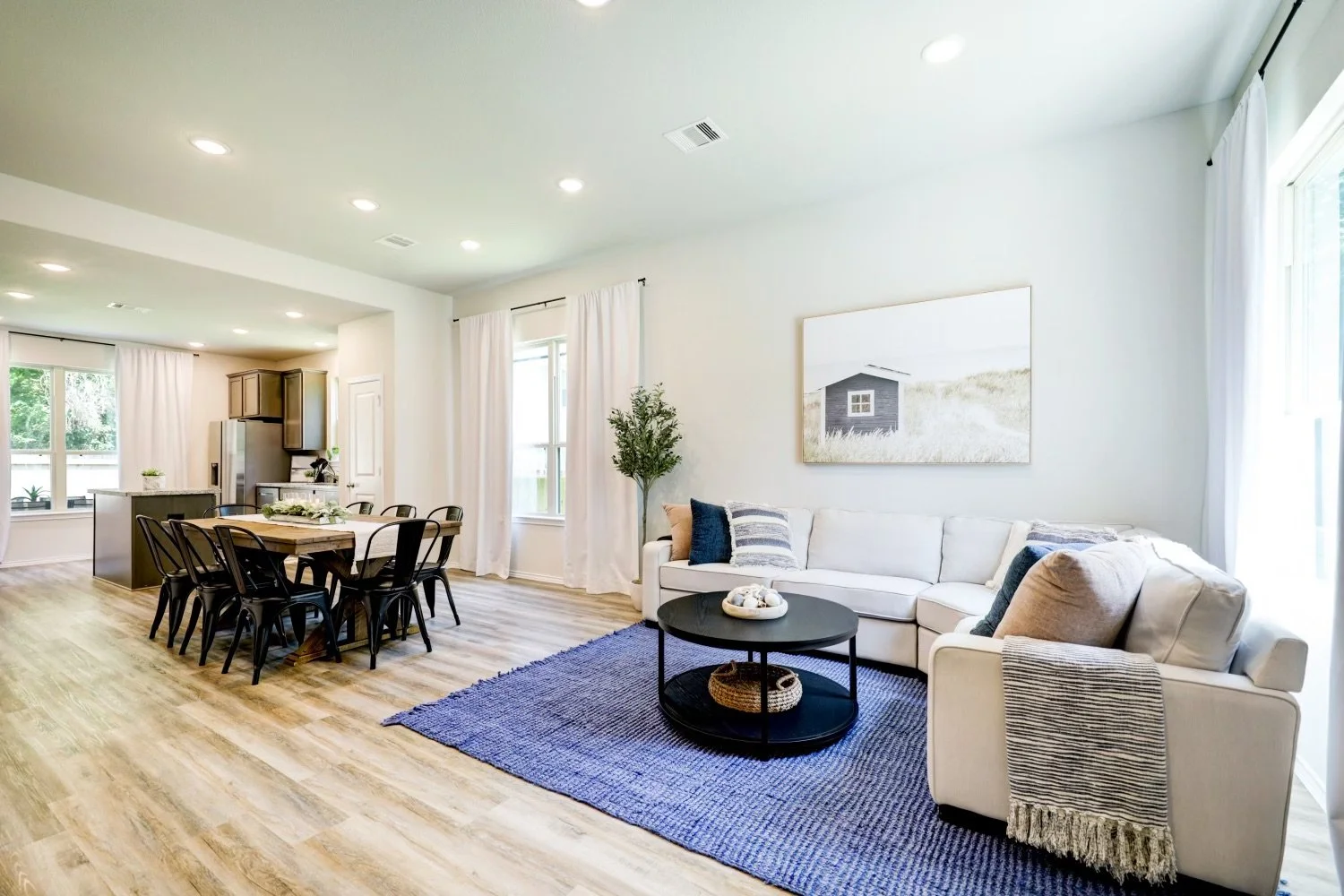 Bright modern open-concept living room and kitchen with white sectional sofa, blue rug, dining table, and large windows