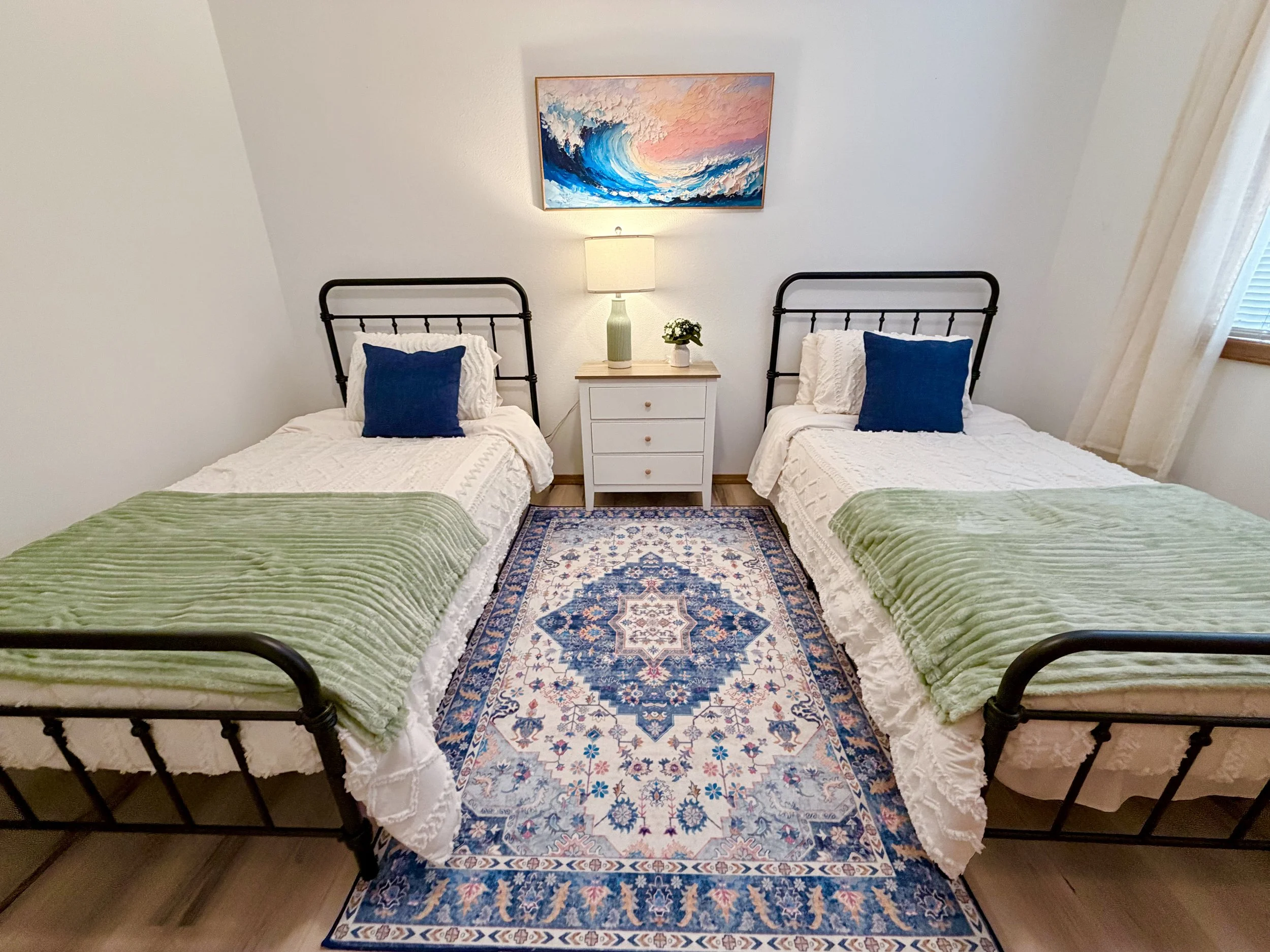 Bright bedroom with two twin iron beds, green blankets, blue pillows, a white nightstand, ocean wave artwork, and patterned area rug