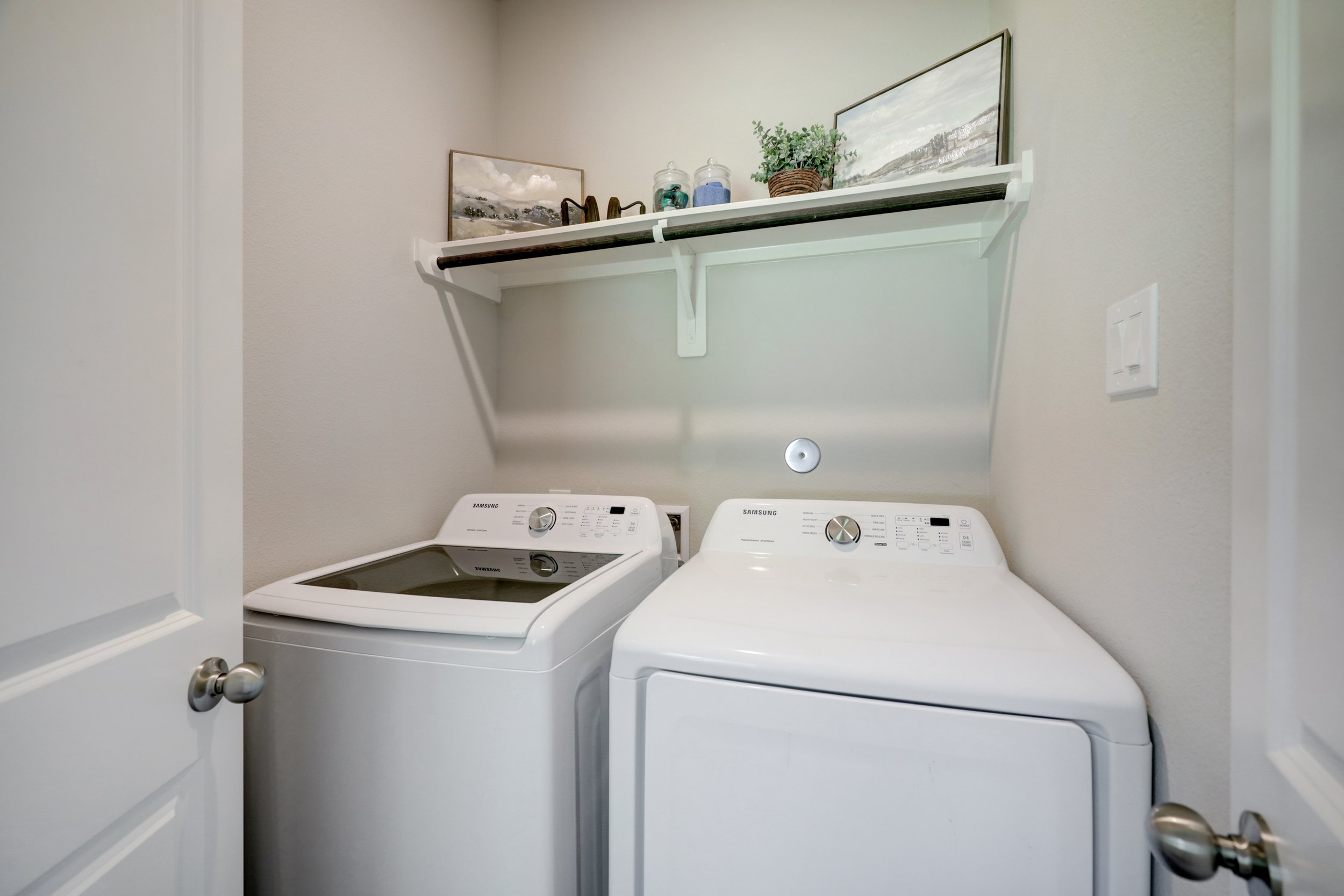 Laundry room with white Samsung washer and dryer beneath a decorative shelf