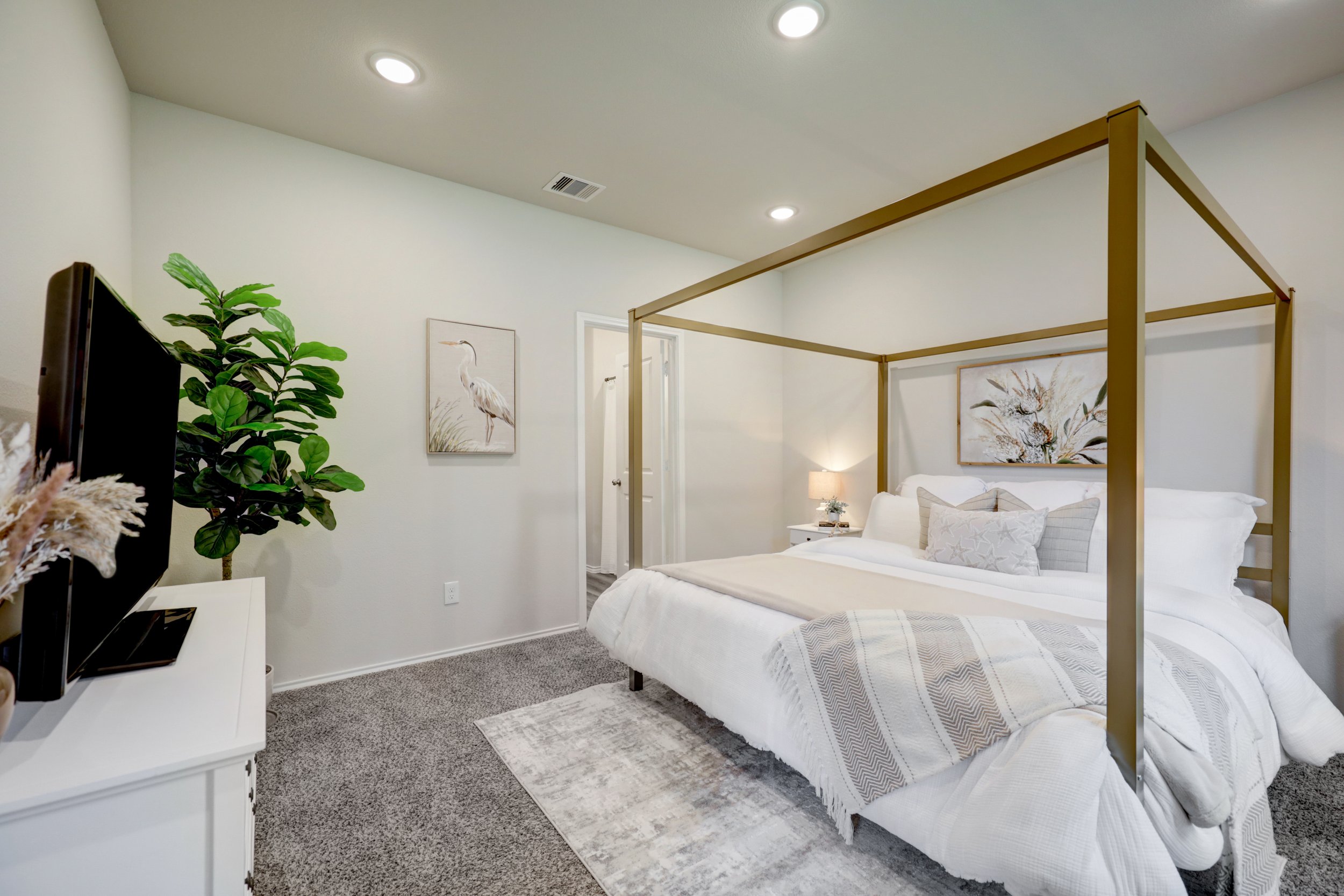 Bright modern bedroom with a gold four-poster bed, white bedding, wall art, television, and potted plant.