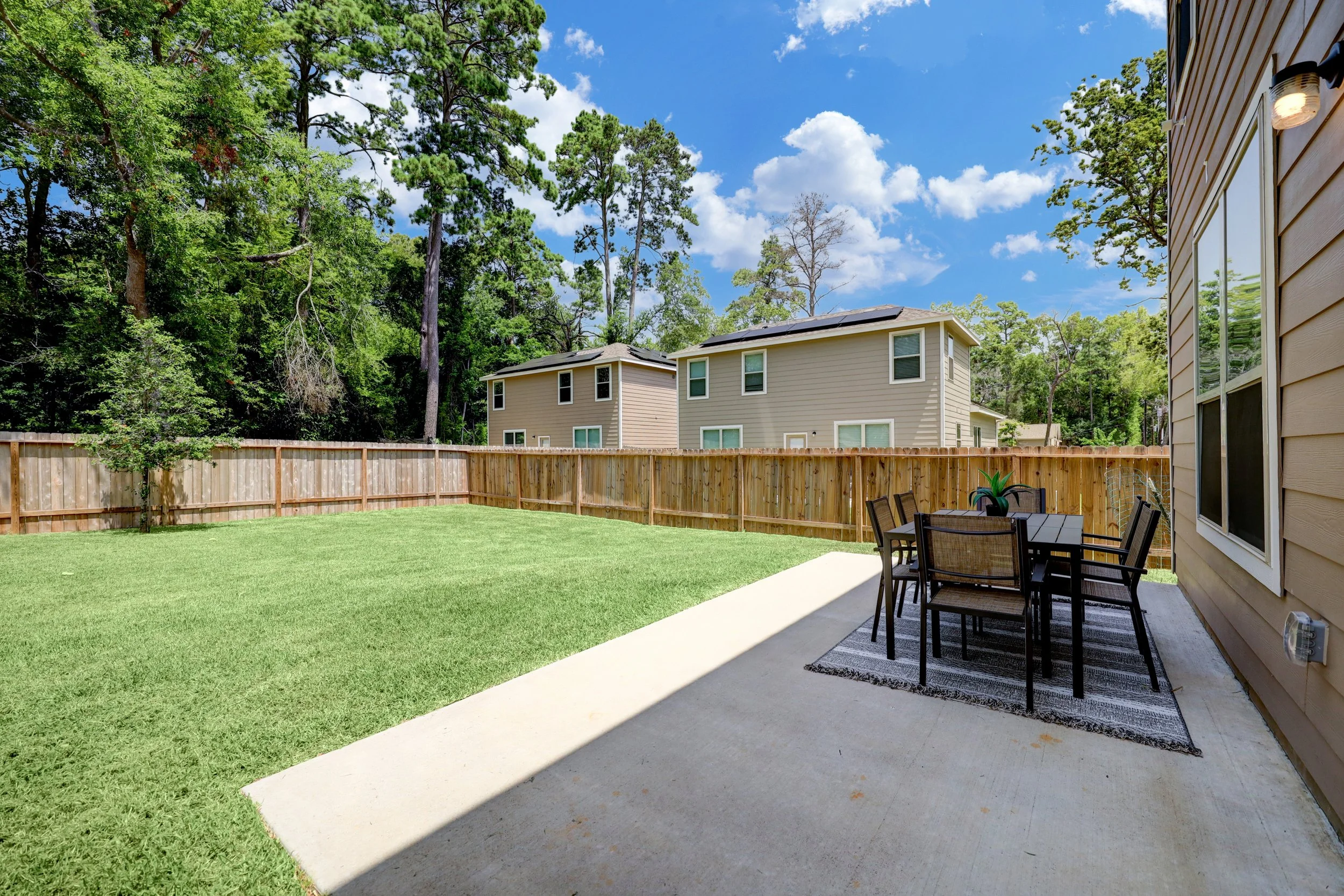 Fenced backyard with a lush lawn, concrete patio, outdoor dining set, and neighboring homes beneath a sunny blue sky