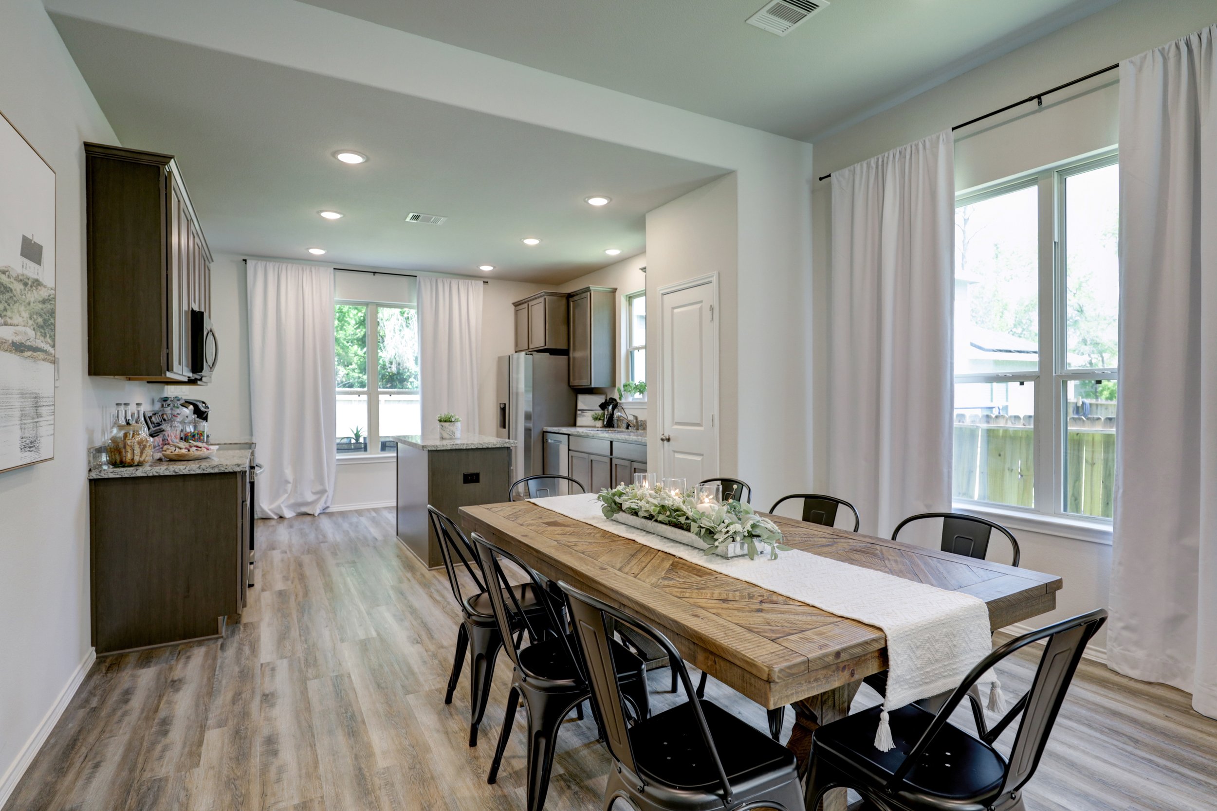 Bright open-concept kitchen and dining room with dark wood cabinets, granite countertops, island, large farmhouse dining table, black chairs, and white-curtained windows.
