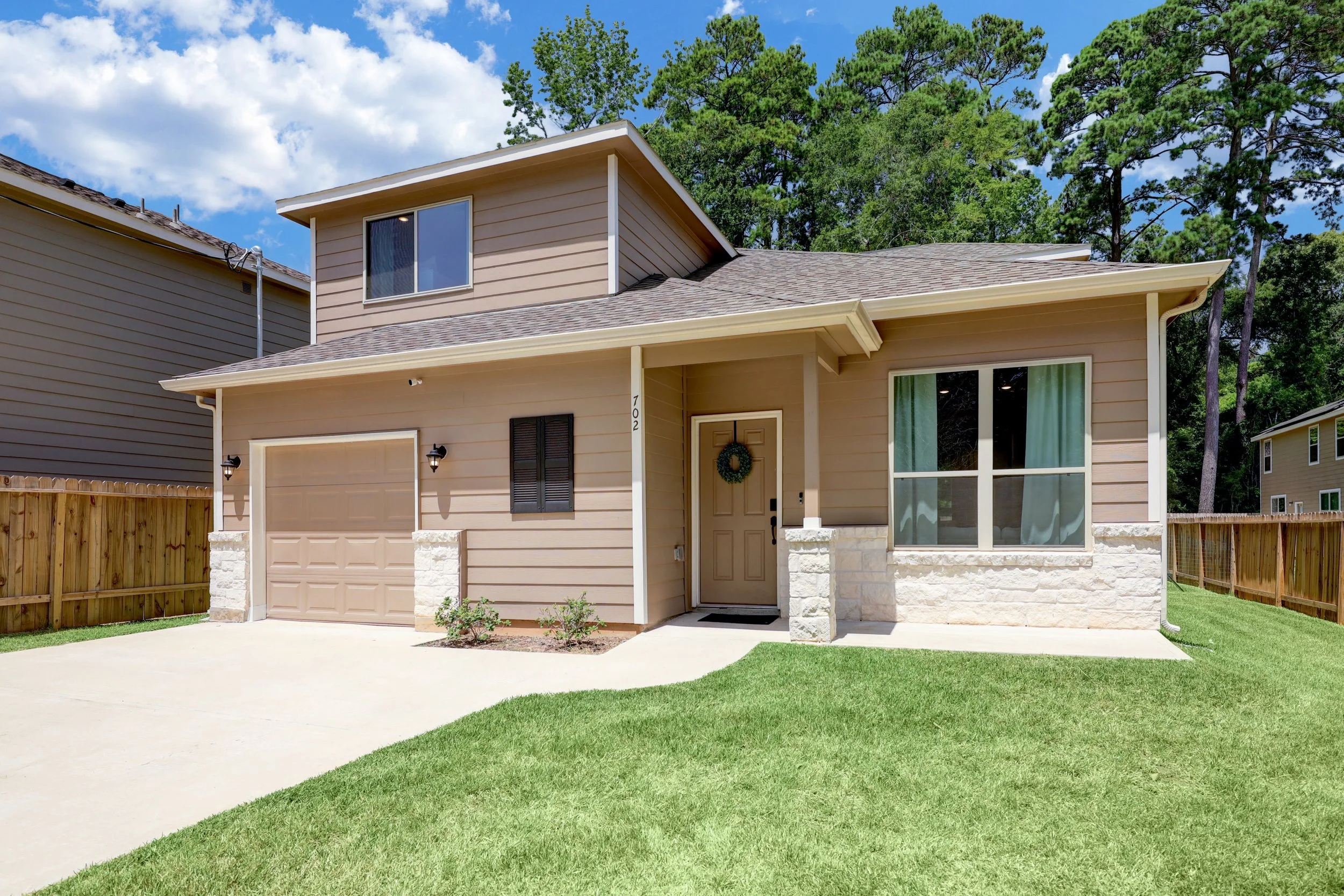 Exterior view of a tan two-story suburban home with a garage, covered entryway, fenced yard, and mature pine trees